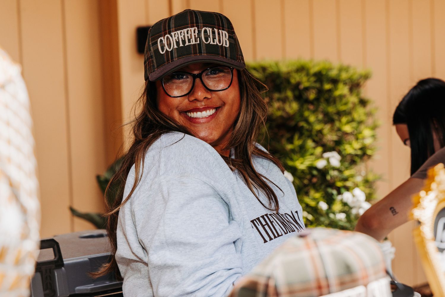 Smiling woman wearing a Coffee Club cap and glasses at an outdoor casual gathering.