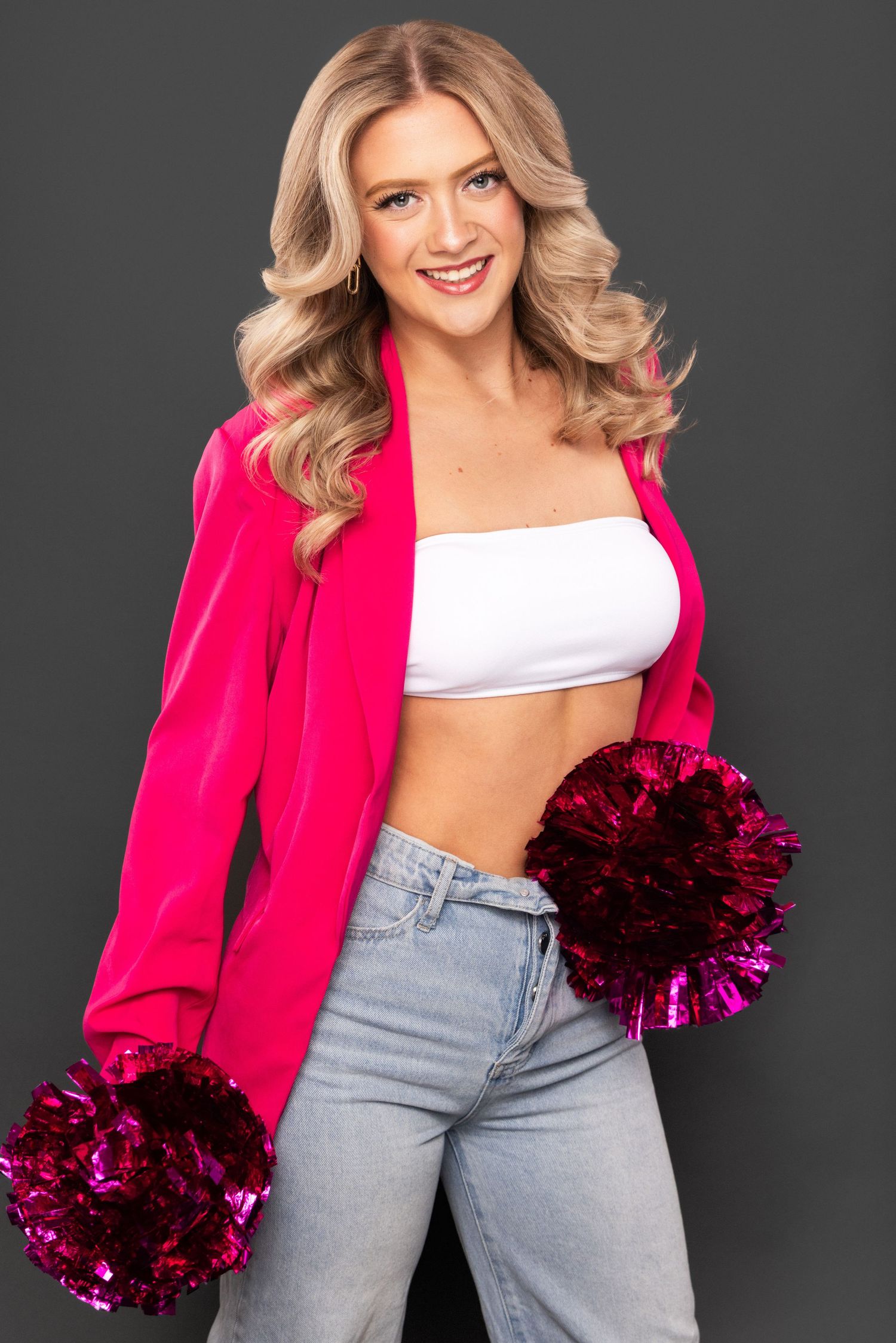 A model in light wash jeans and a hot pink blazer poses with magenta pom poms against a dark background.