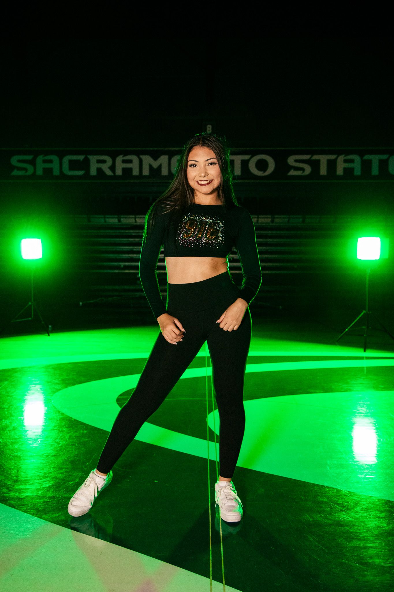 Dance performer in black outfit and white sneakers poses under dramatic green spotlights.