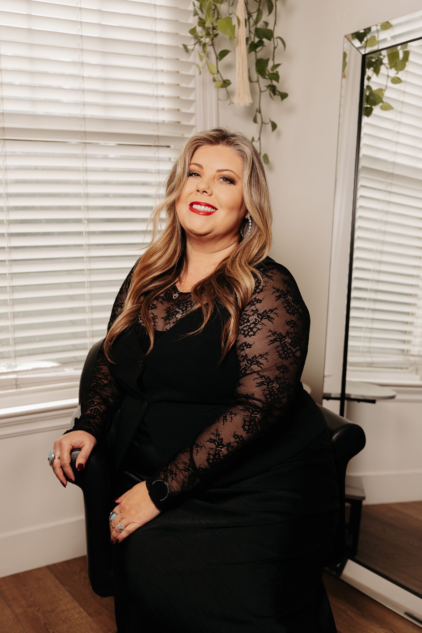 A person wearing a black lace-sleeved dress sits in a chair near window blinds and hanging plants.
