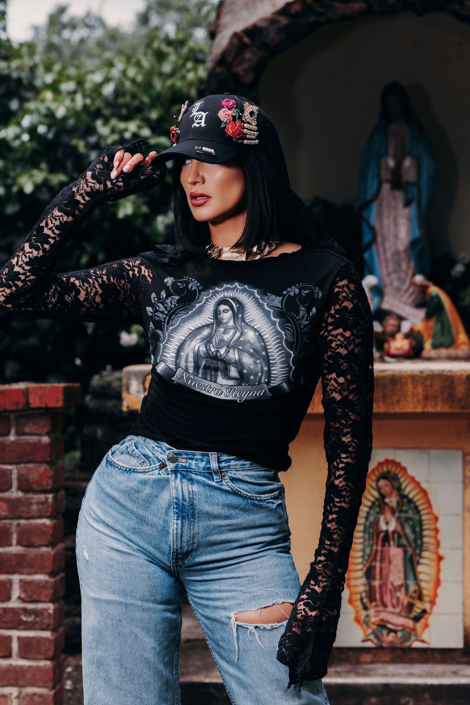 Woman in black lace top with Virgin Mary graphic, ripped jeans, and floral embroidered cap poses outdoors near religious statues.