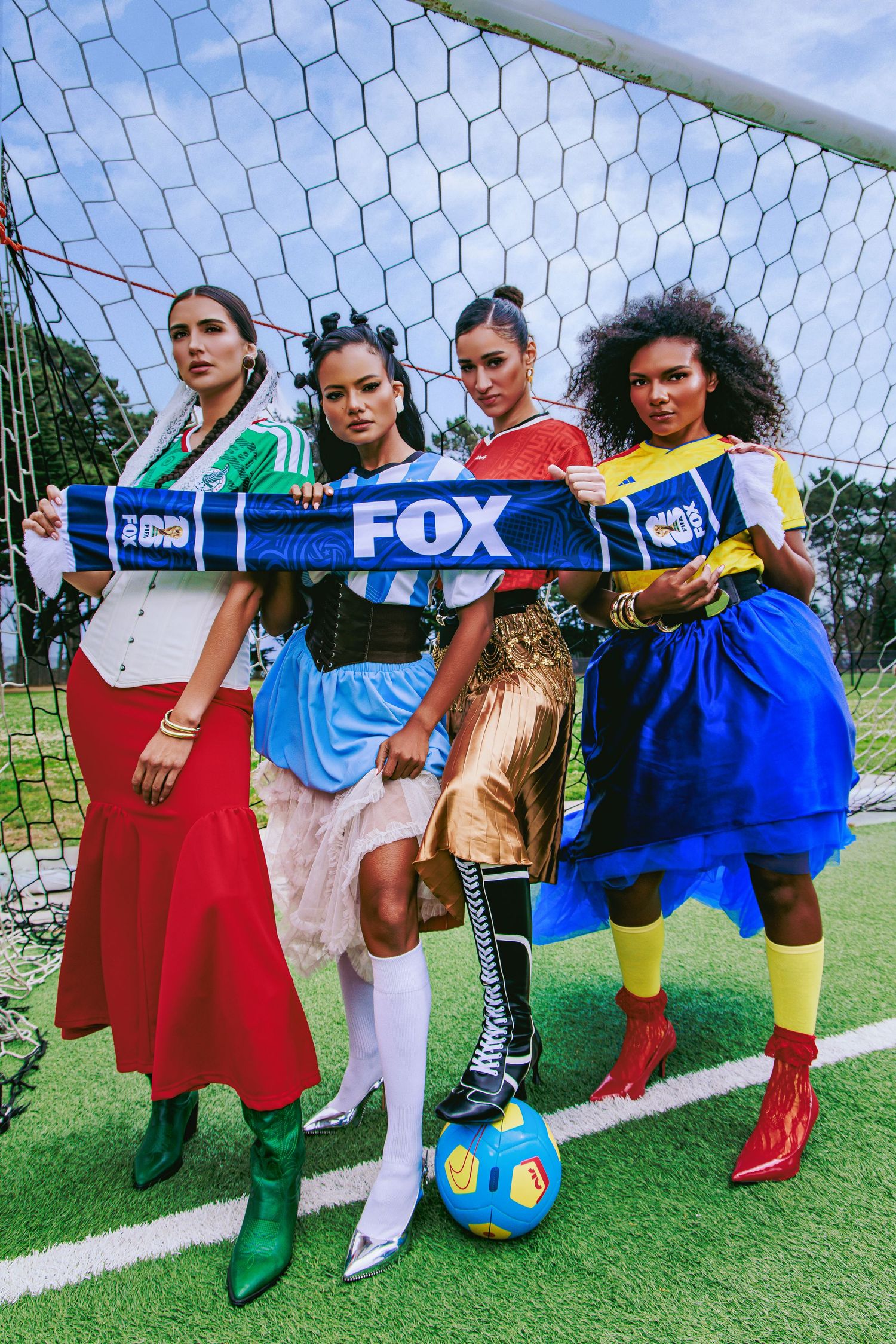 Four stylish women pose at a soccer goal holding a FOX sports scarf, wearing colorful fashion-forward outfits with a soccer ball.