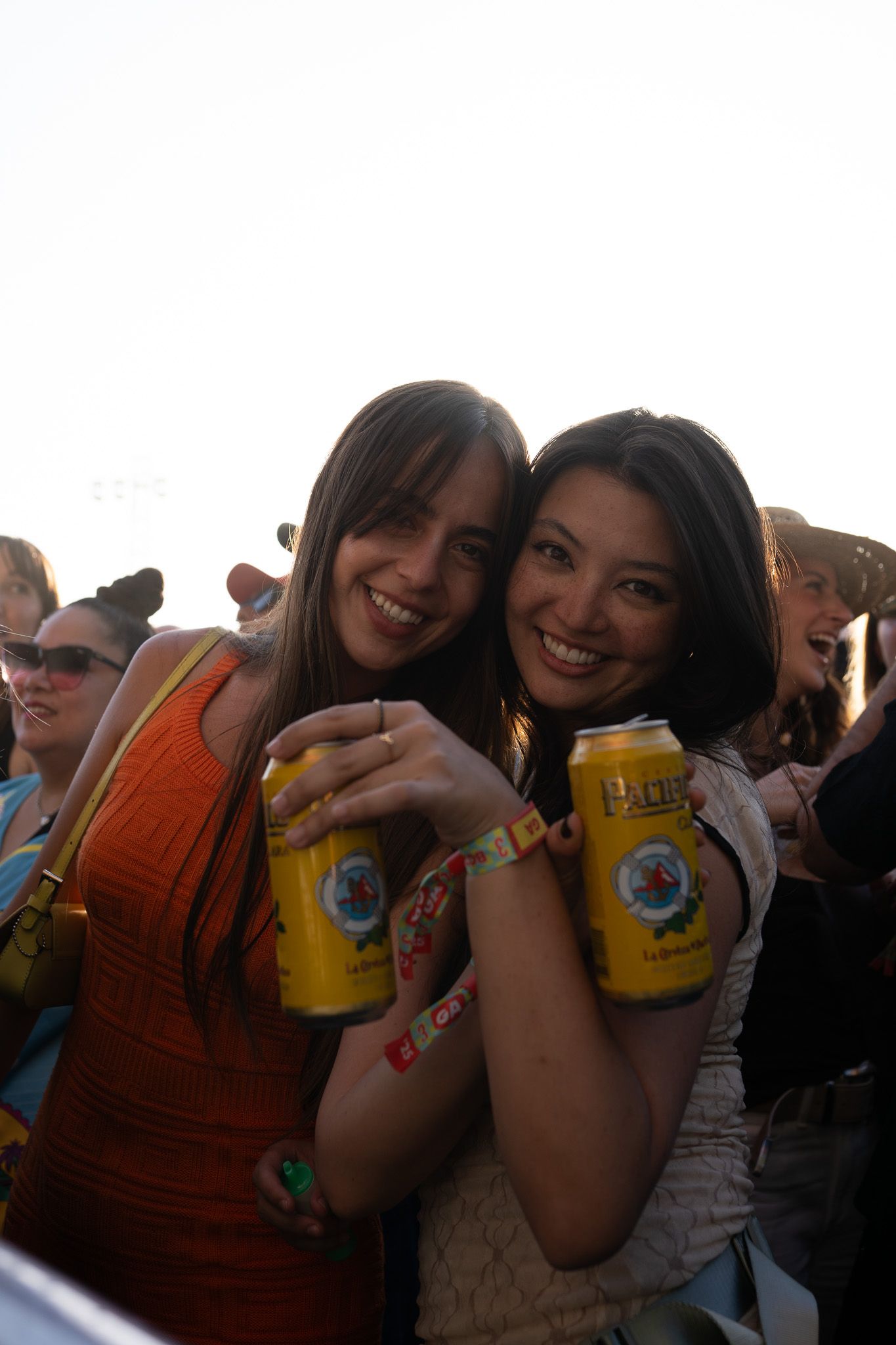 Two smiling women holding yellow beer cans at an outdoor festival, posing together in a crowd.