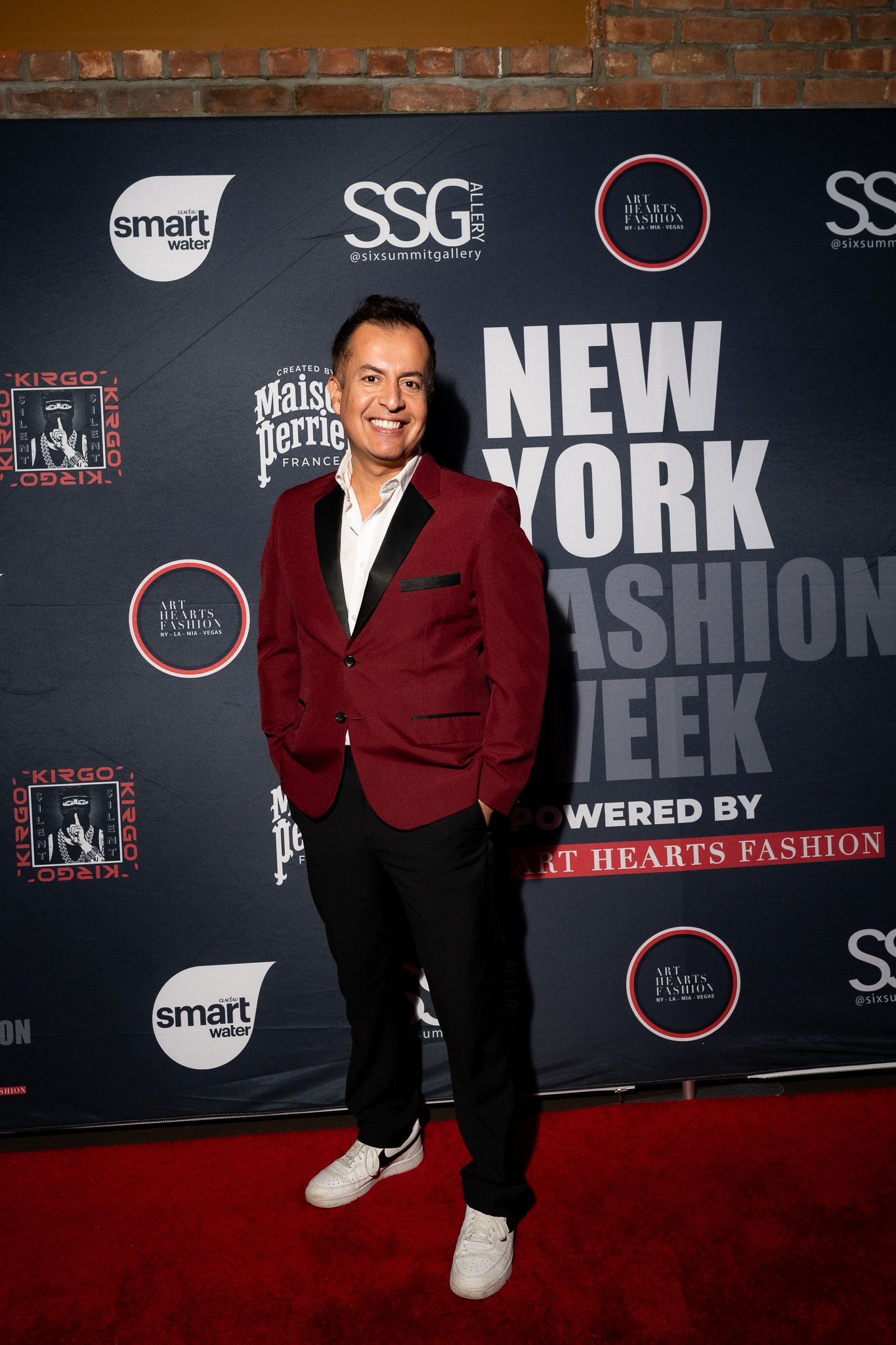 A person in a burgundy velvet blazer and black pants poses against a dark backdrop with fashion week event signage.