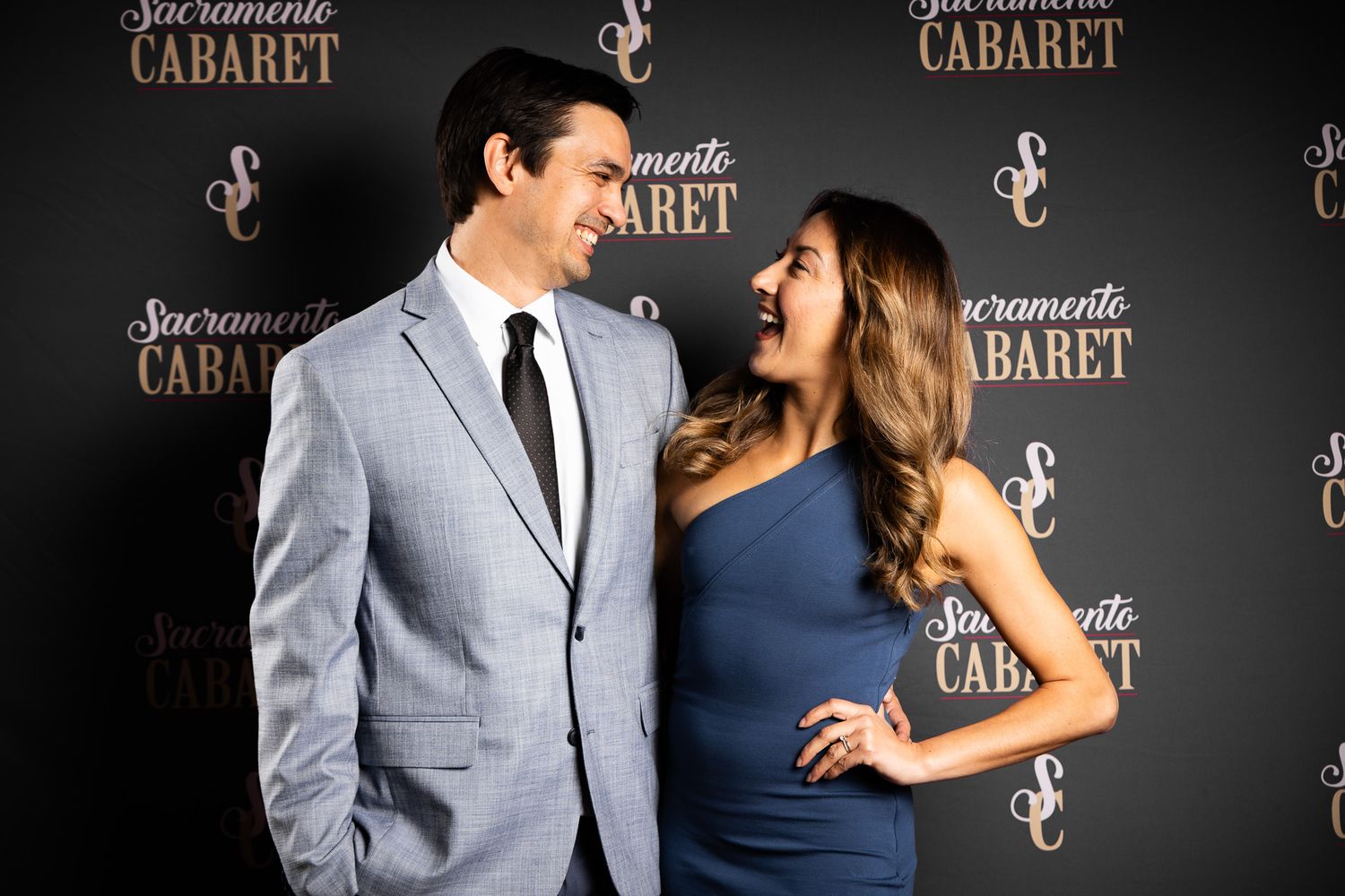 Elegant couple poses at a cabaret event backdrop, man in light gray suit and woman in navy blue dress.