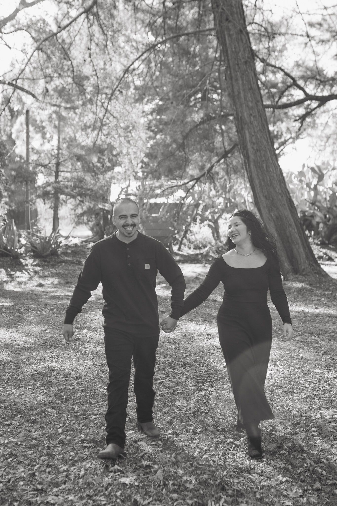 A couple in black clothing walks together through a wooded area in a series of candid black and white photos.