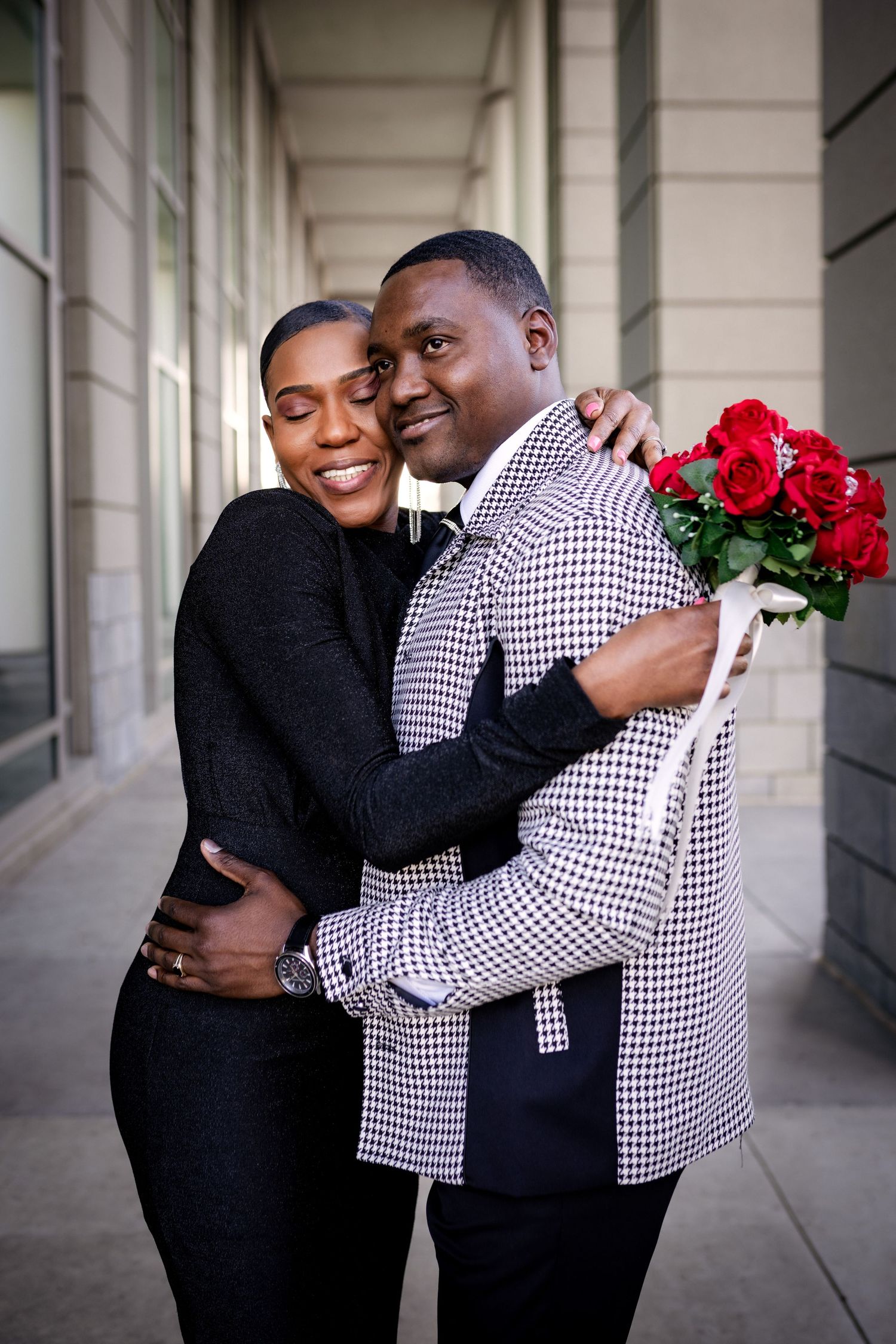 A couple embraces in formal attire while holding red roses in an elegant columned hallway.