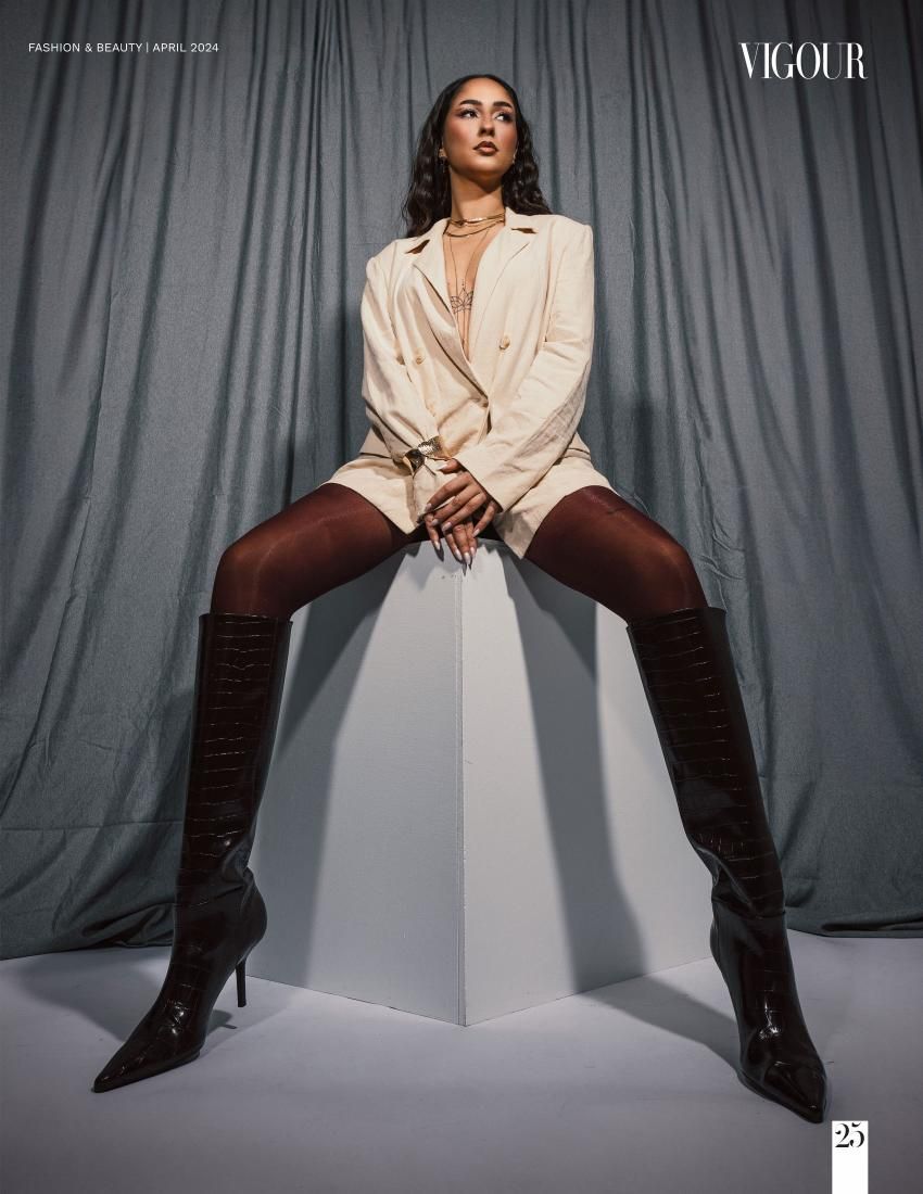 Fashion model in beige blazer and thigh-high boots sits confidently on white cube against gray backdrop.