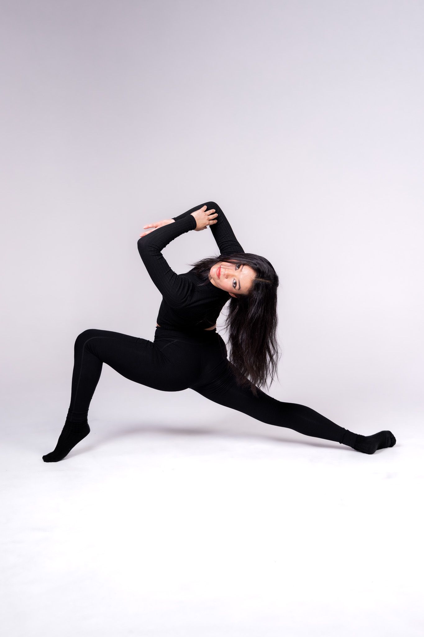 Dancer in black outfit performs dramatic split pose on light background.