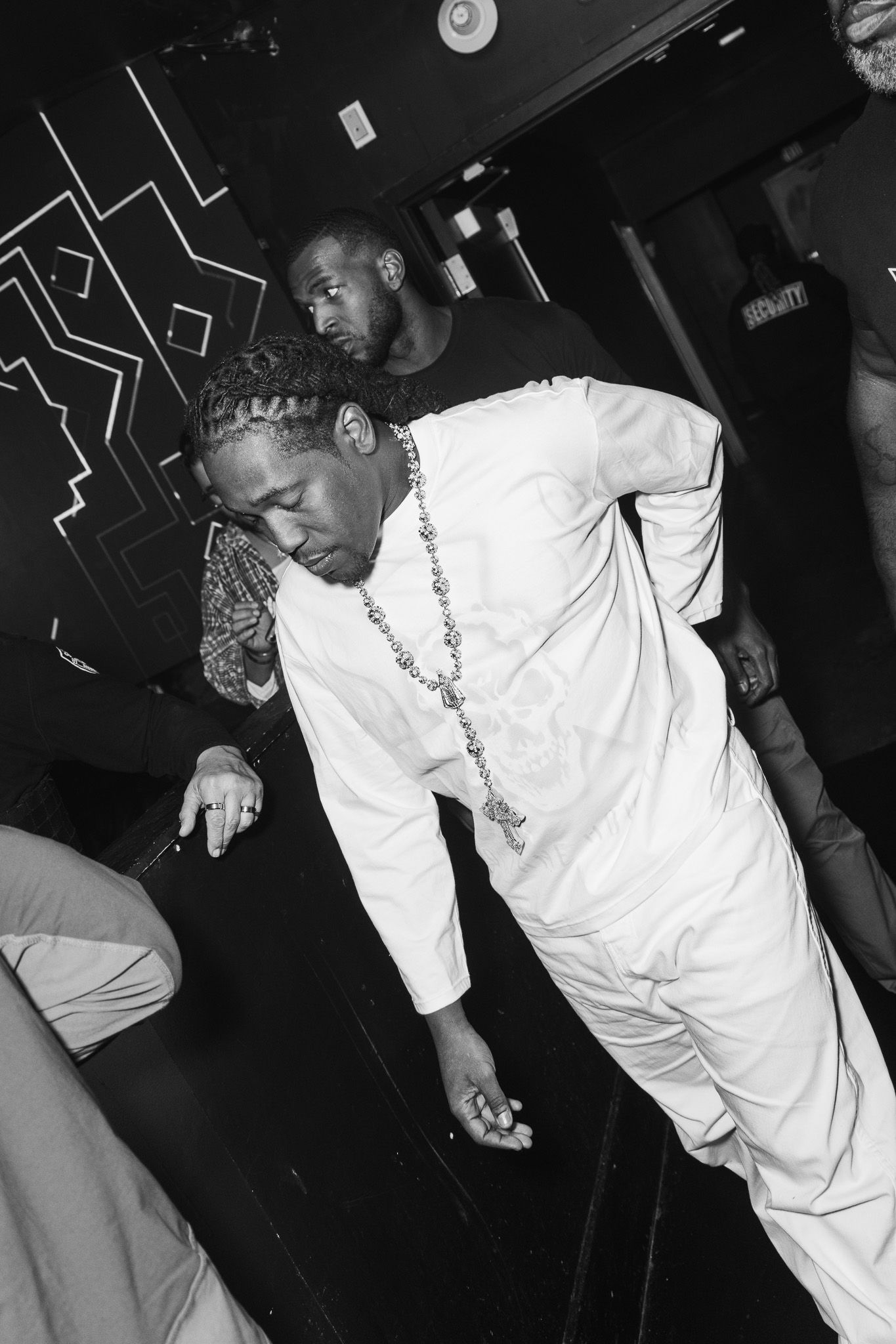 Black and white photo of a man in white outfit and chain necklace leaning forward in a nightclub setting.