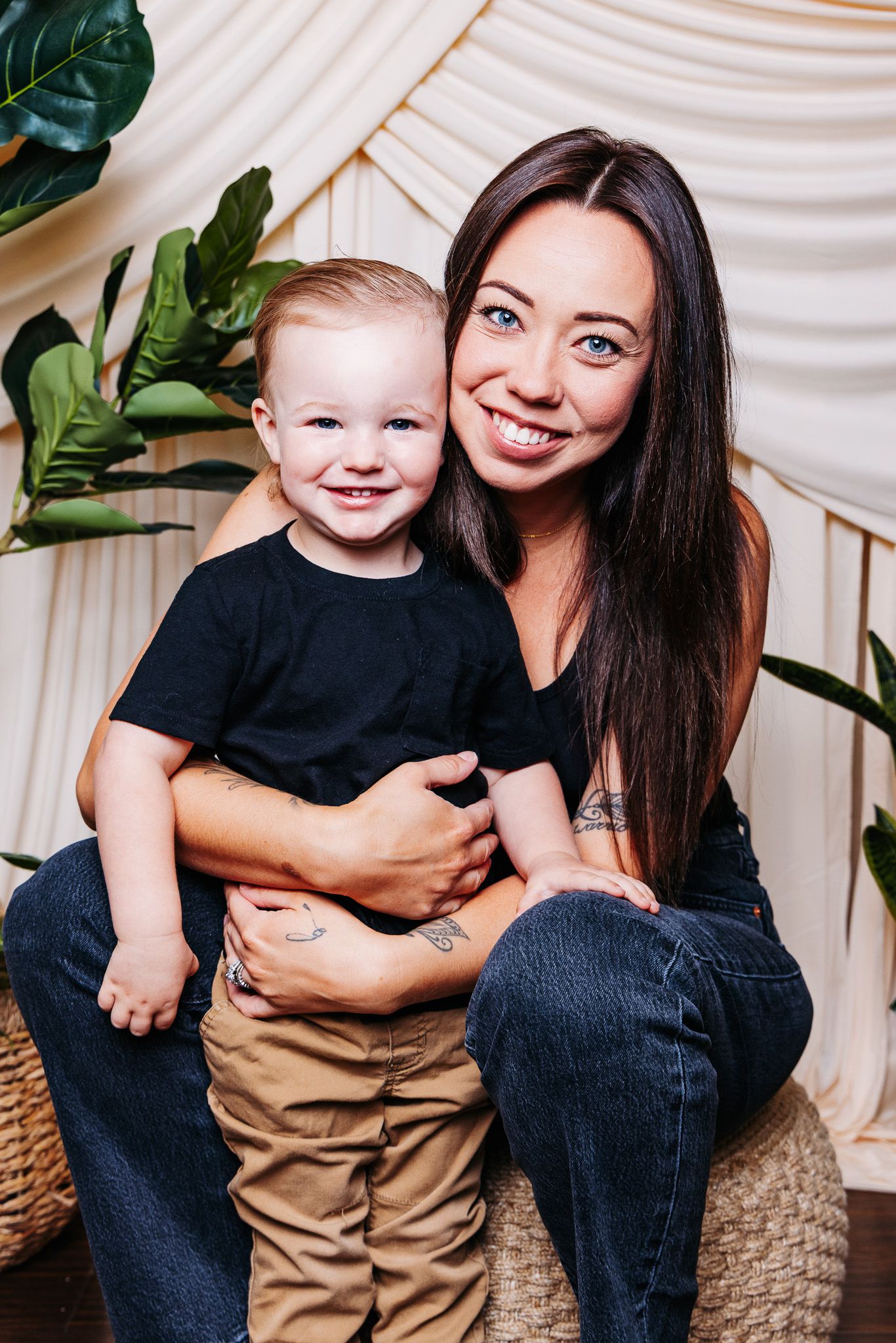 A smiling woman with dark hair holds a happy toddler boy, both wearing black shirts, posed before green plants.