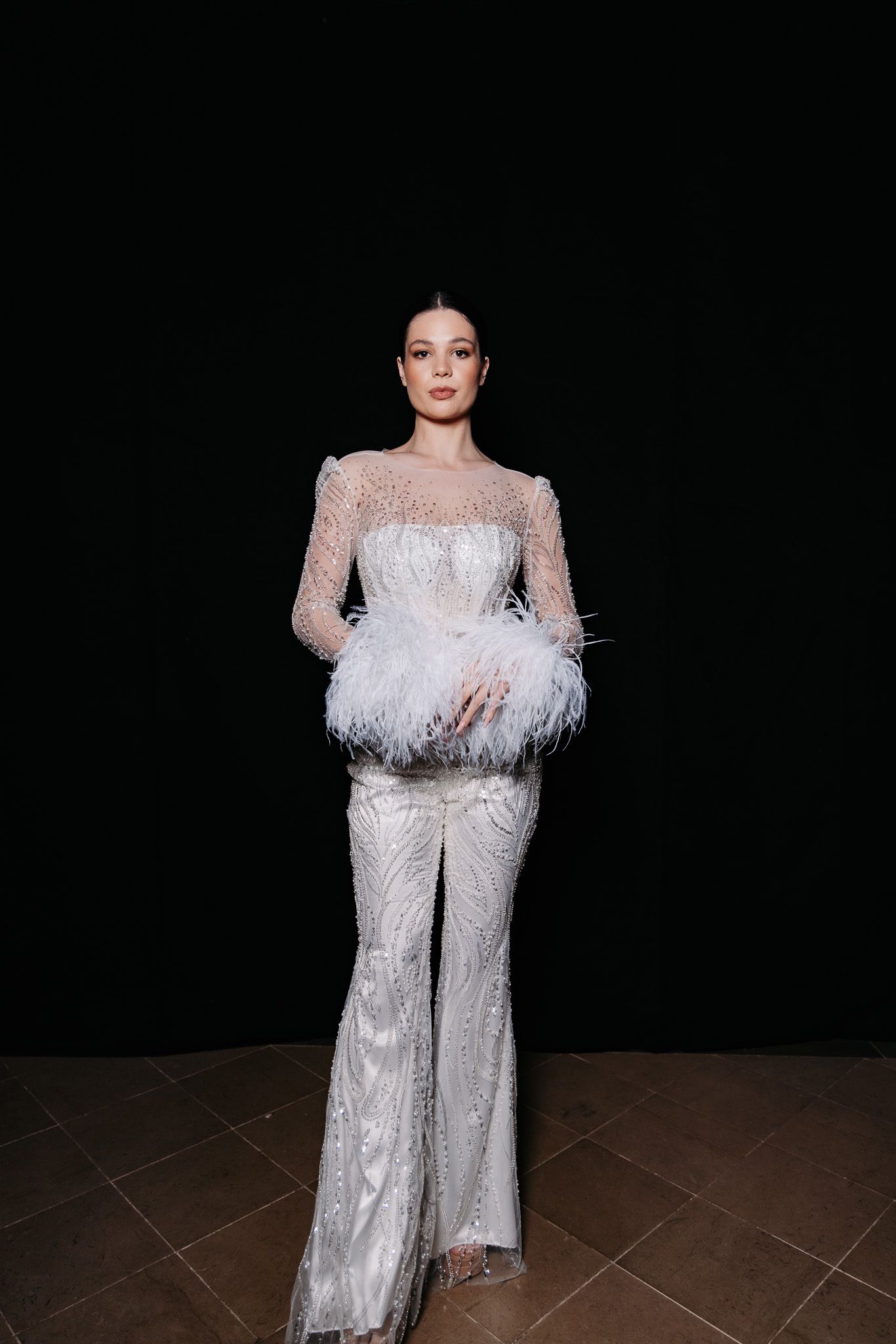 Model walks runway in glamorous white sequined gown with feather details and dramatic long sleeves.