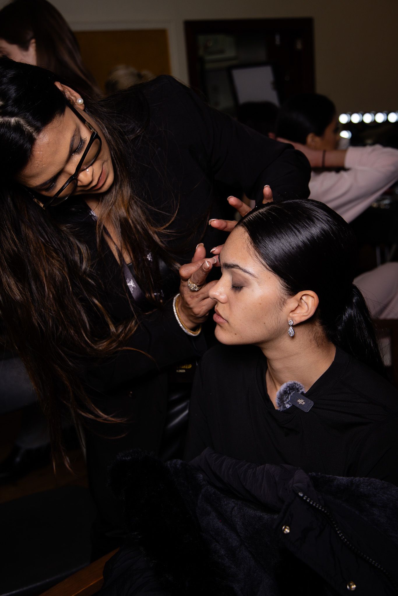 Darkly lit backstage scene showing makeup artists preparing models for runway show.
