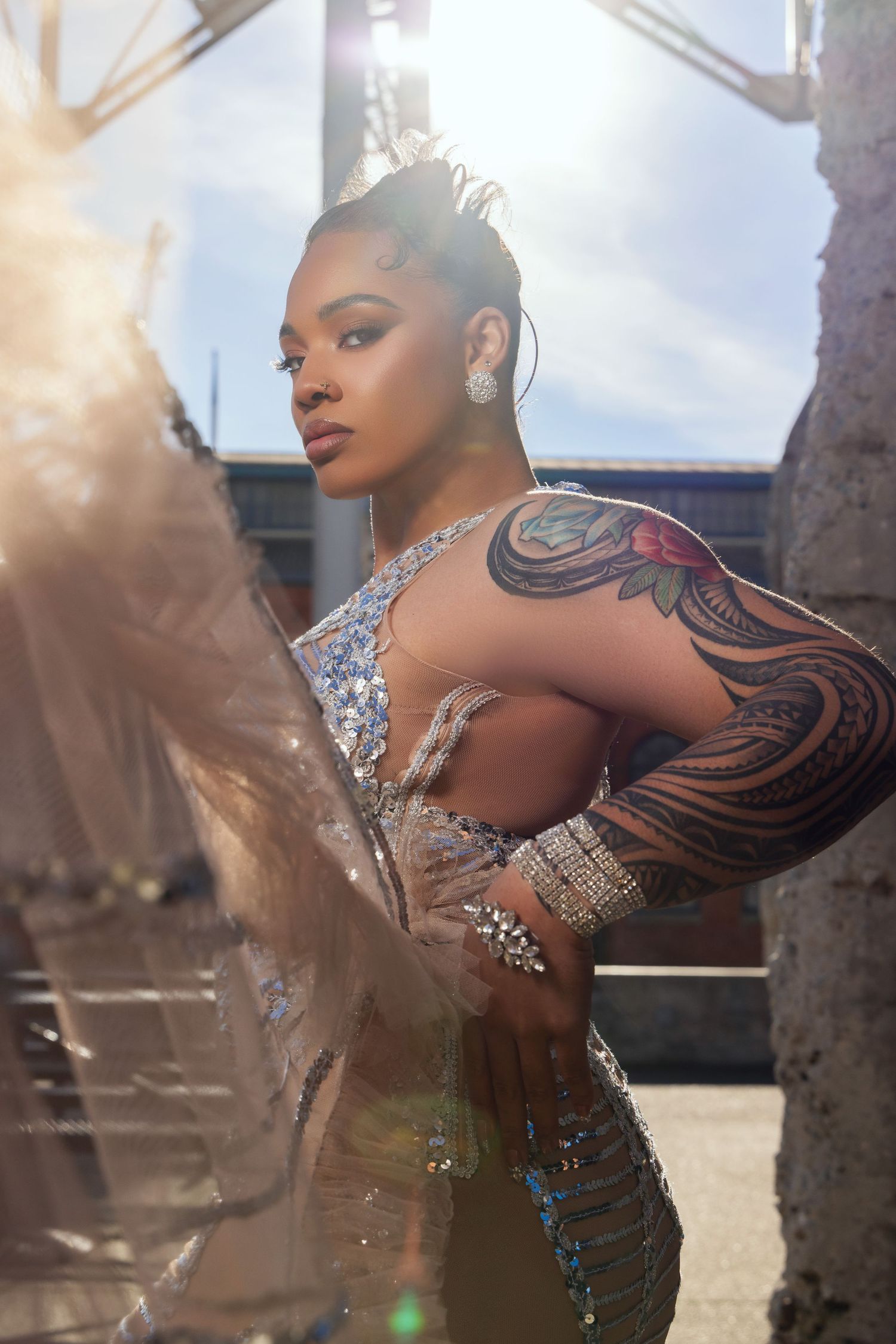 A glamorous model in a sparkly dress with intricate tattoo sleeve poses outdoors in dramatic sunlit photography.