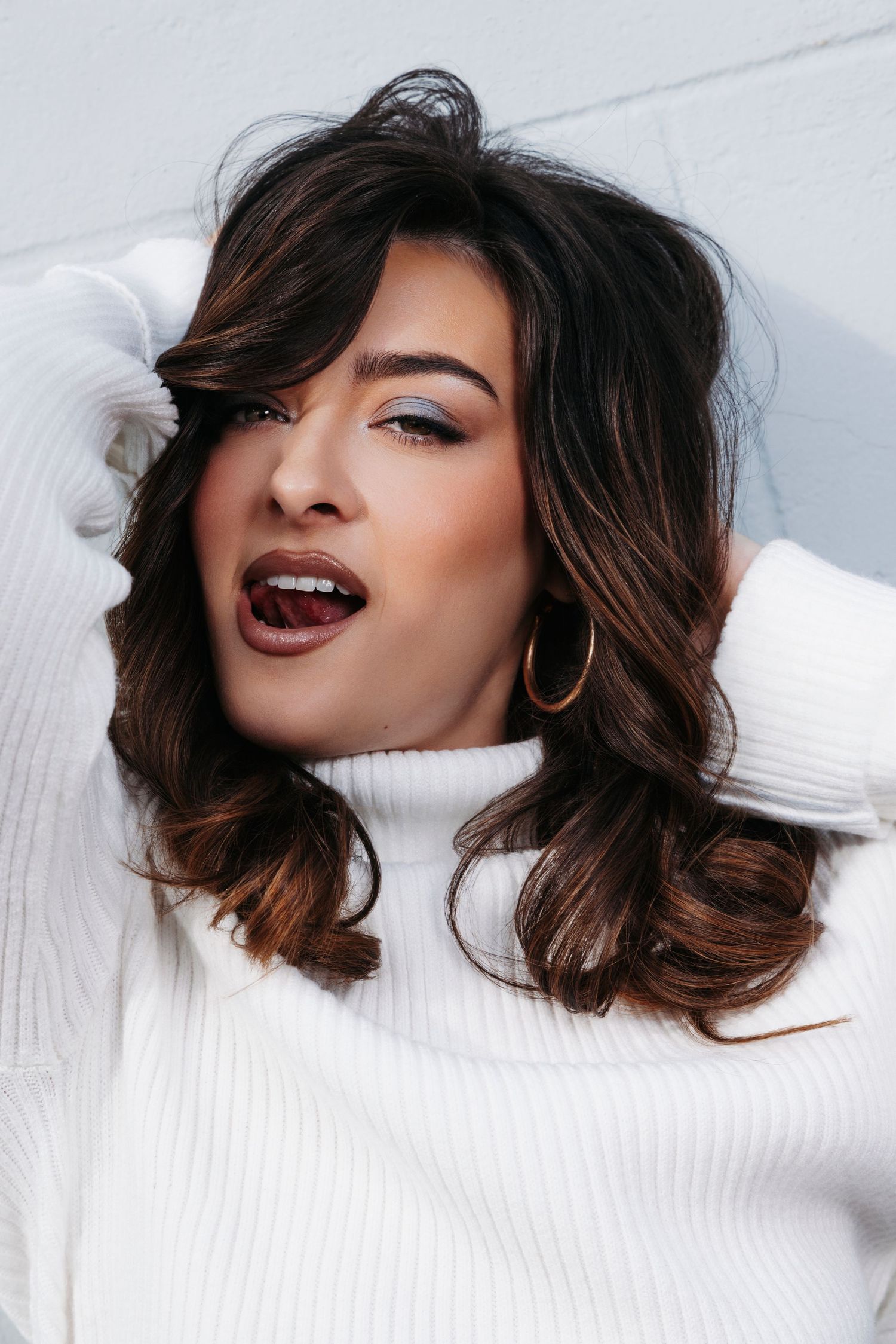 Portrait of someone with wavy brown hair wearing a white sweater and gold hoop earrings against a light background.