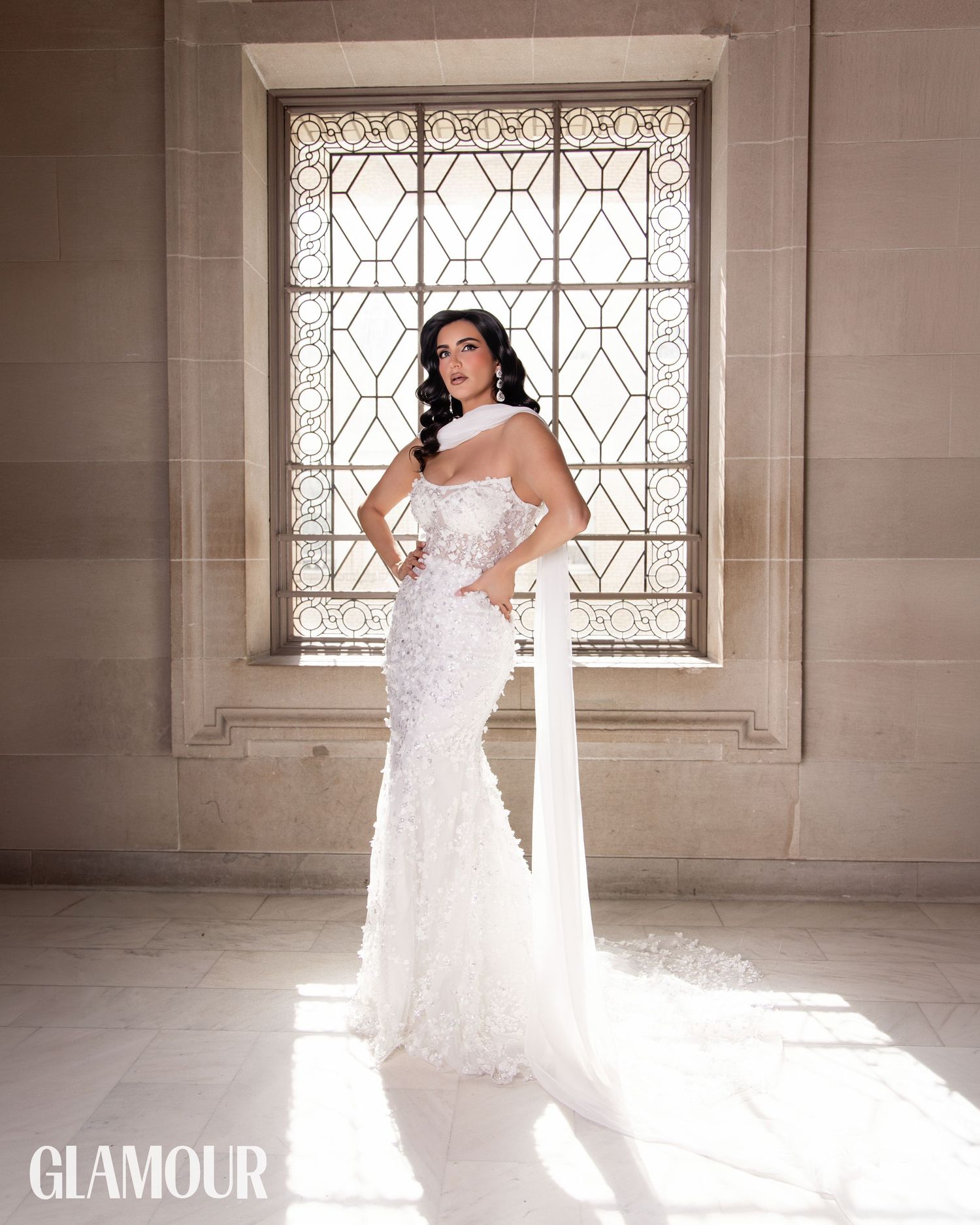 A bride in a strapless white lace wedding gown poses elegantly in front of an ornate window with natural light streaming in.
