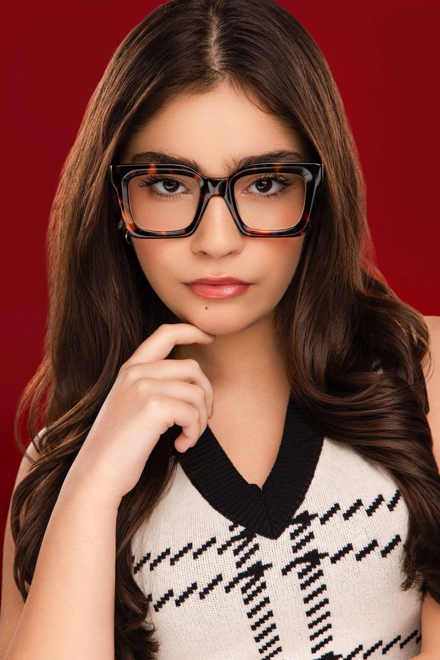 Studio portrait against red backdrop featuring black rimmed glasses and black and white patterned sweater.