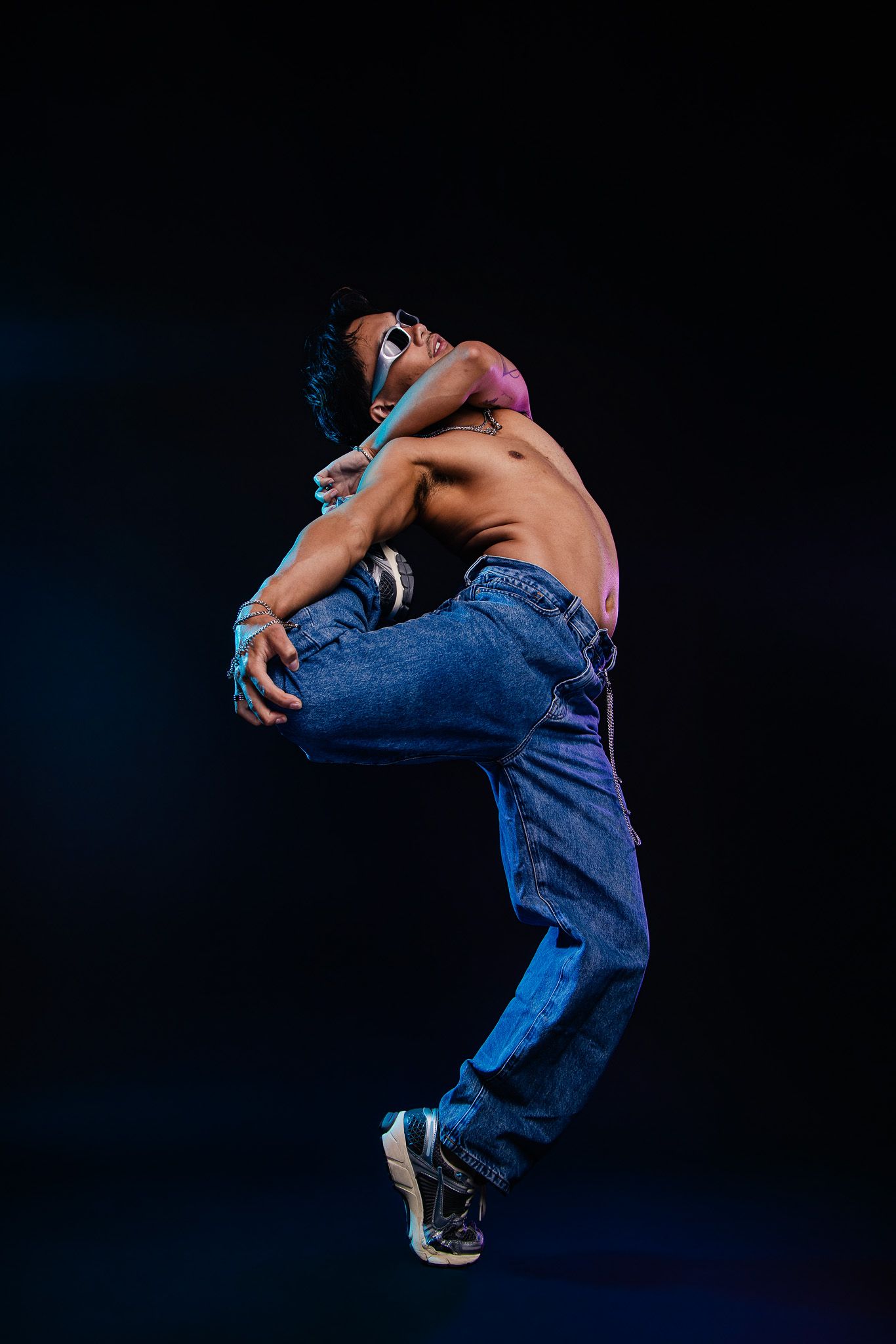 Shirtless male dancer mid-jump wearing jeans and sunglasses against a dark background.