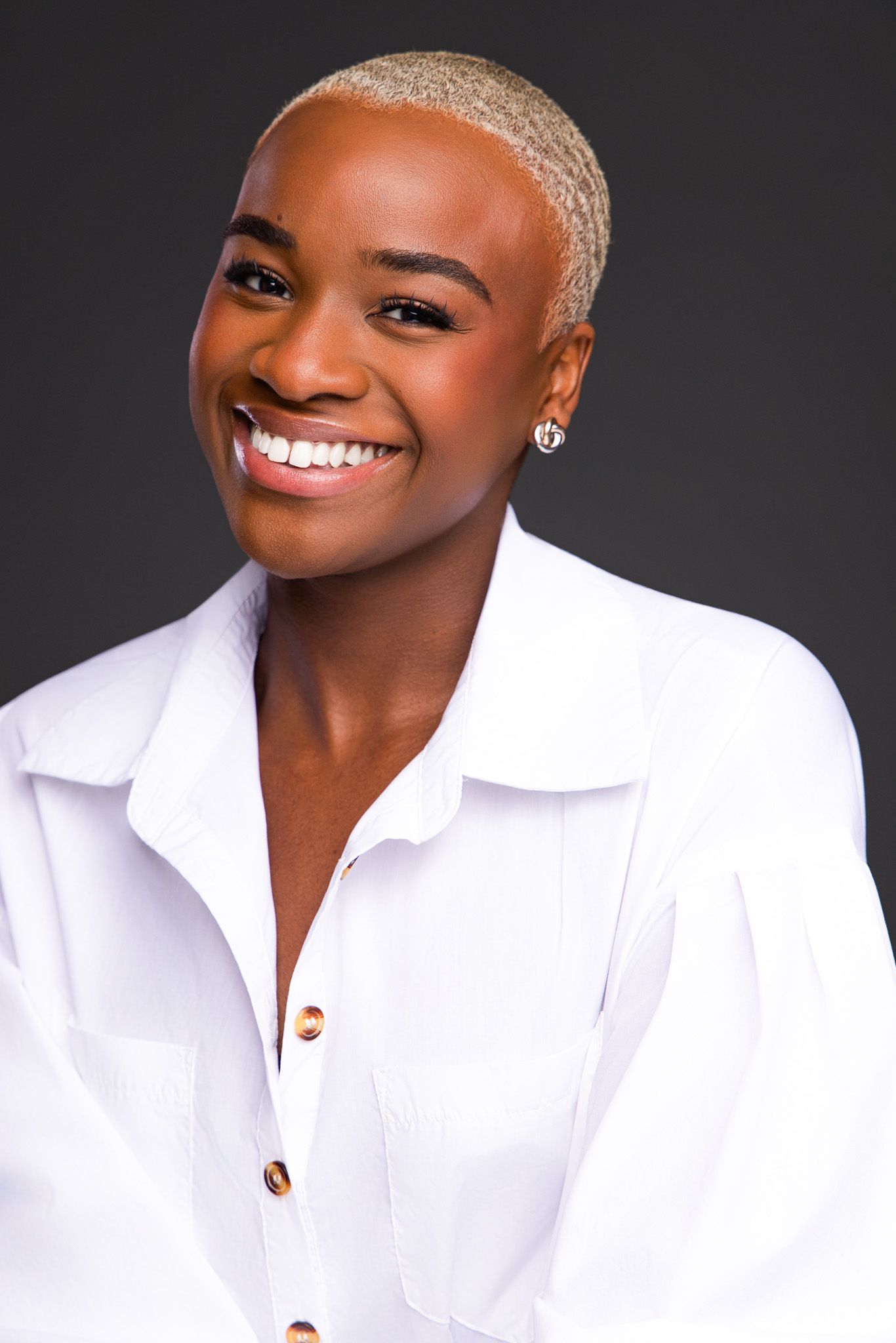 Smiling Black woman with short blonde hair wearing a white button-up shirt, posing against a dark gray background.