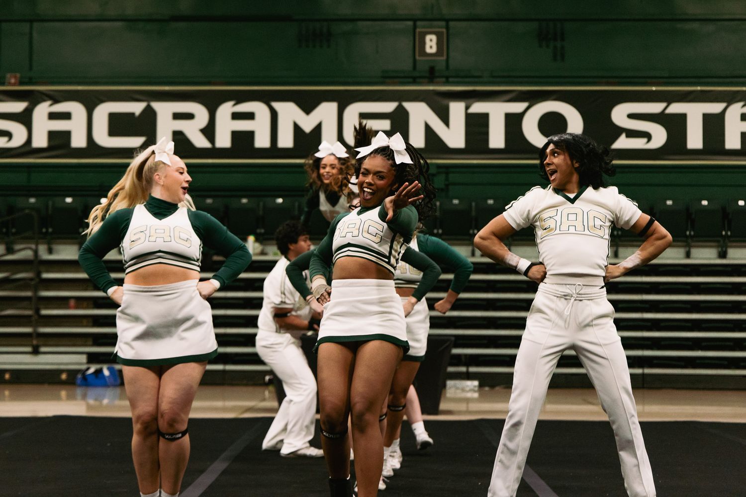 Sacramento State cheerleaders perform in green and white uniforms on a gymnasium floor during a spirited routine.