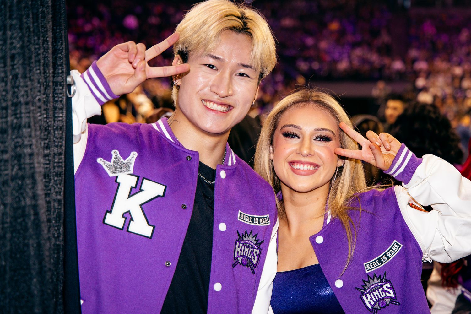 Two young people wearing purple Sacramento Kings varsity jackets pose together making peace signs at an indoor event.