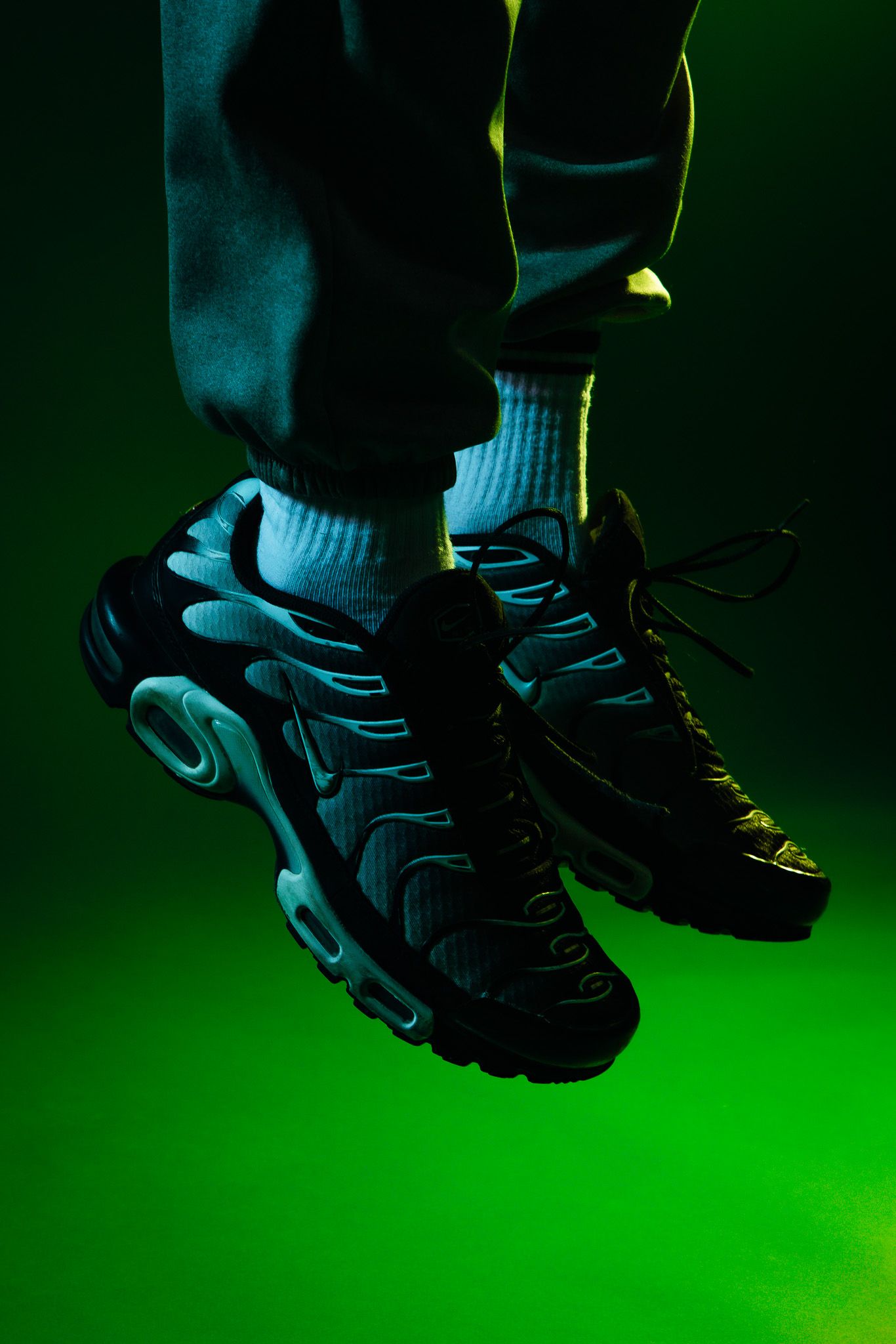 Nike Air Max Plus sneakers in black and teal on a green background, worn with white socks and dark joggers.