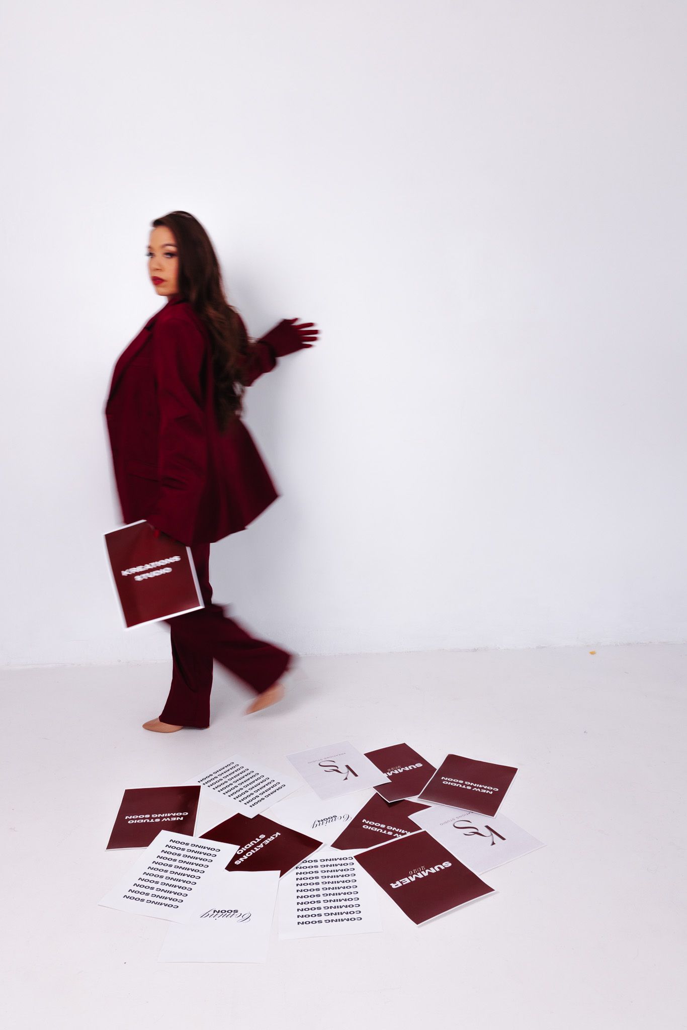Woman in red blazer walking away from scattered white and burgundy papers on white studio floor.