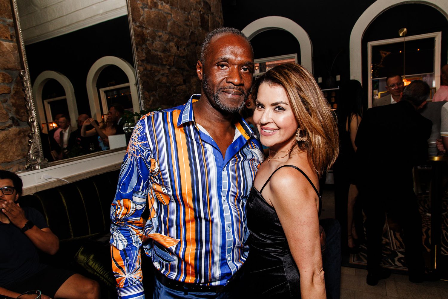 A man in a colorful striped shirt poses with a woman in a black dress at an indoor social event.