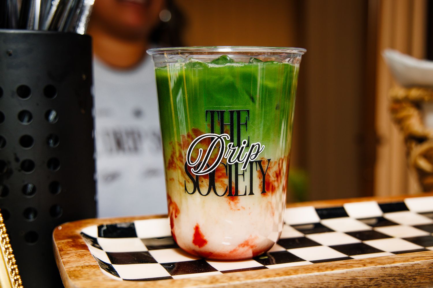 Colorful layered green and red drink in a The Drip Society branded cup on a checkered surface.