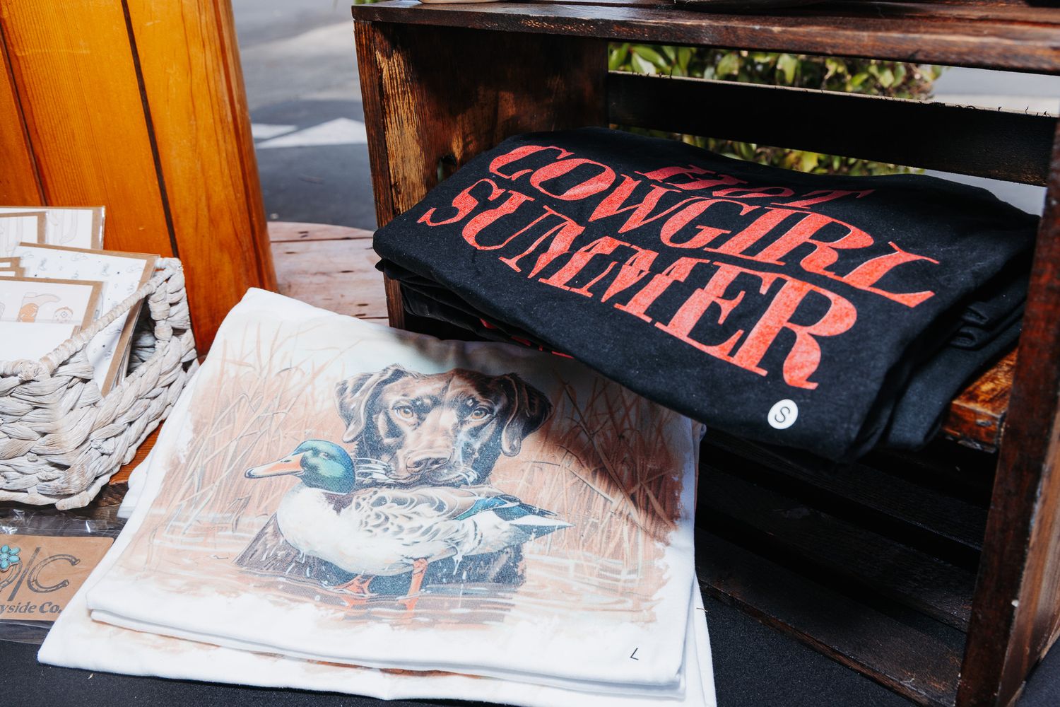 Cowgirl Summer graphic tee displayed in rustic wooden crate at outdoor market alongside vintage duck print artwork.