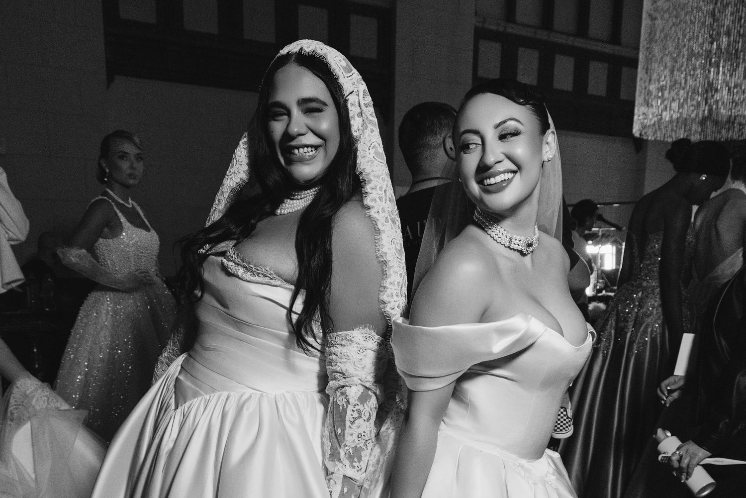 Two women in elegant white gowns smile together at a formal event in a black and white photo.