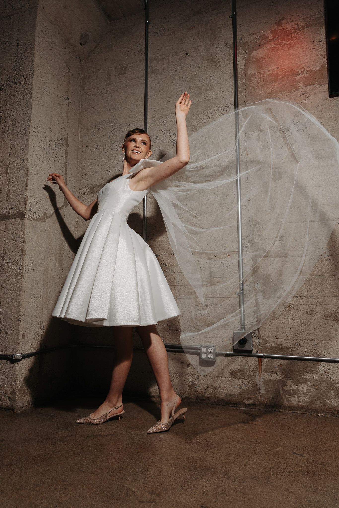 Model in white retro-style tea length wedding dress with flowing veil poses playfully in industrial setting.