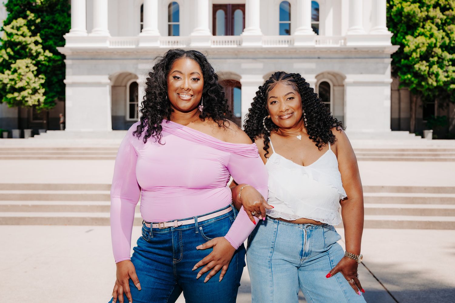 Two people in casual outfits posing and smiling together outdoors in front of a white building.