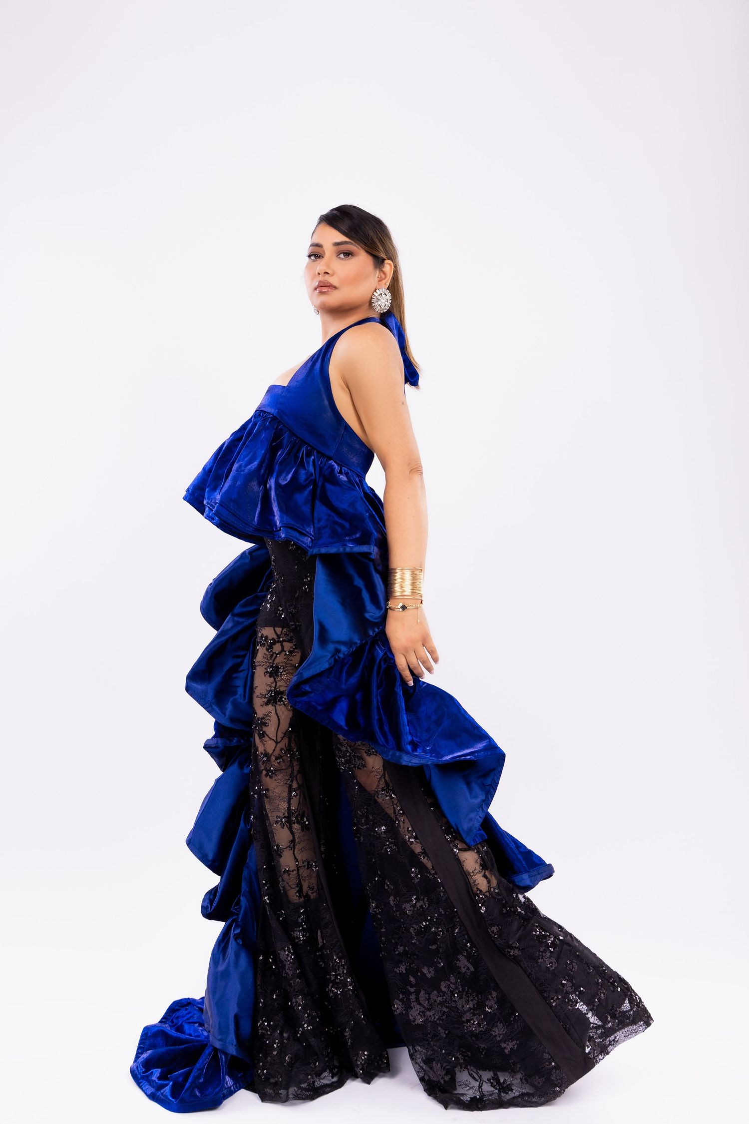 A model poses in a dramatic royal blue and black lace gown with ruffled details against a white background.