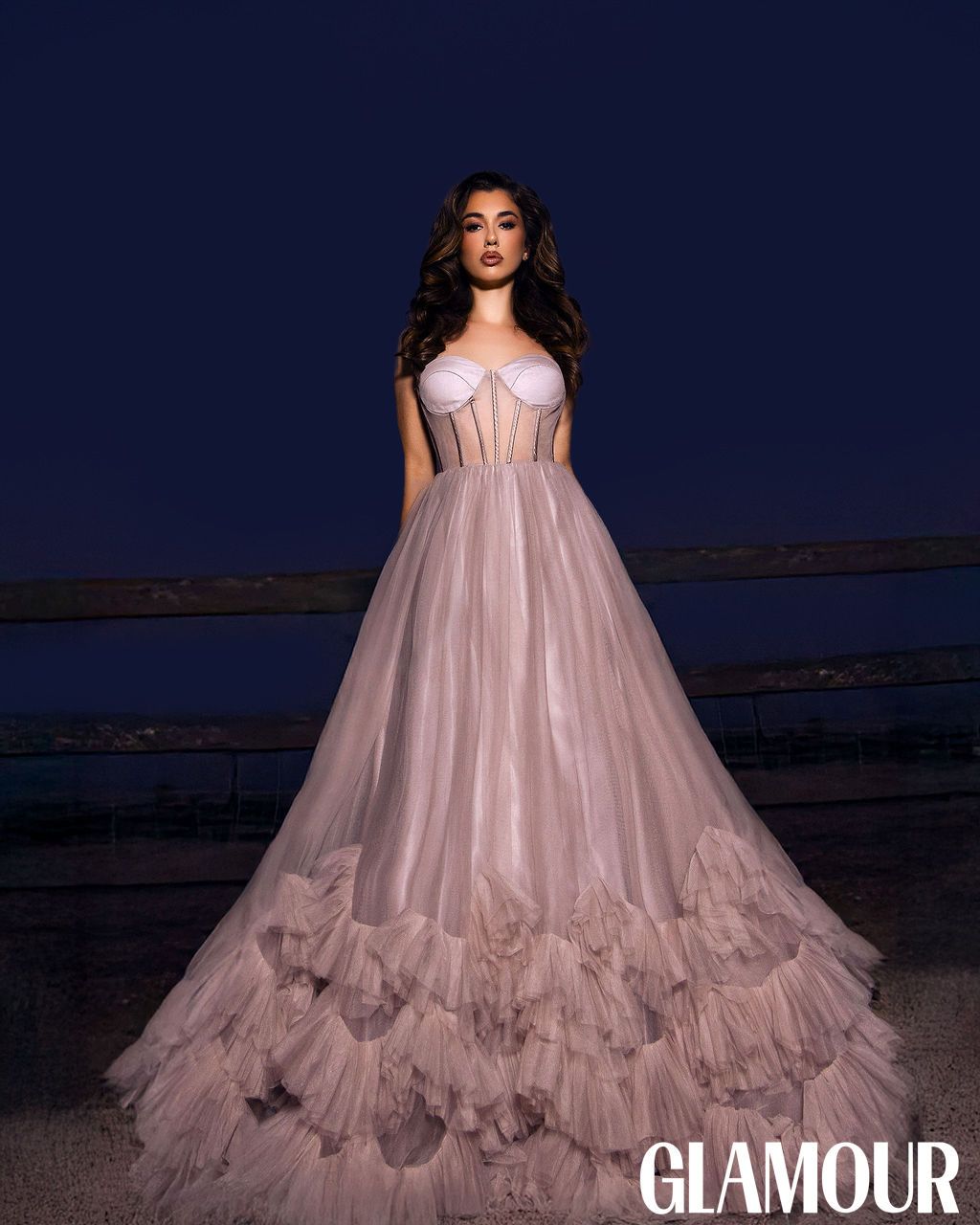 A model in a flowing pink tulle gown poses against a dramatic dark blue evening sky at dusk.