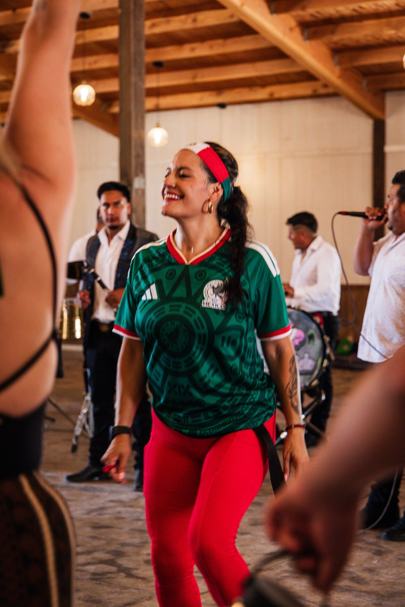 Woman in green Mexico soccer jersey and red leggings smiling and dancing at a lively indoor party.