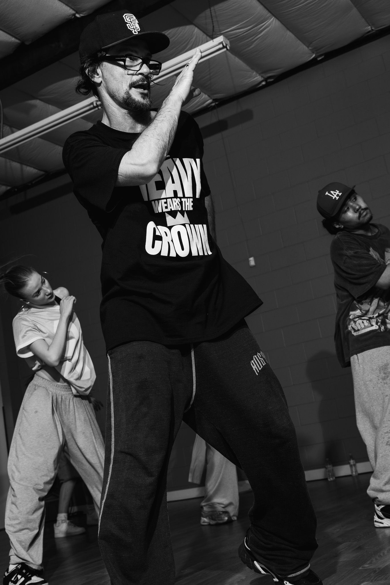 Black and white photo of a man in a SF Giants cap and crown t-shirt dancing in a studio with others.