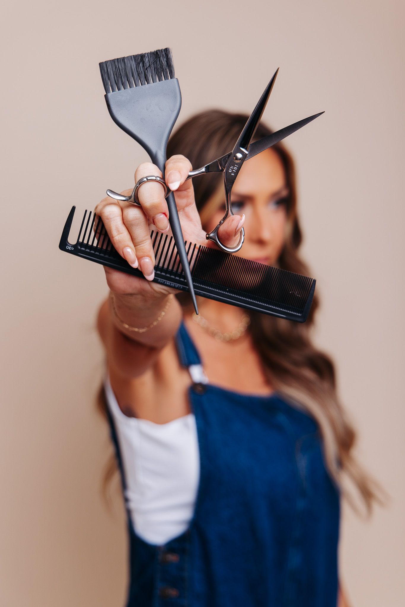 Female hairstylist holding black scissors, comb, and dye brush toward camera in denim apron.