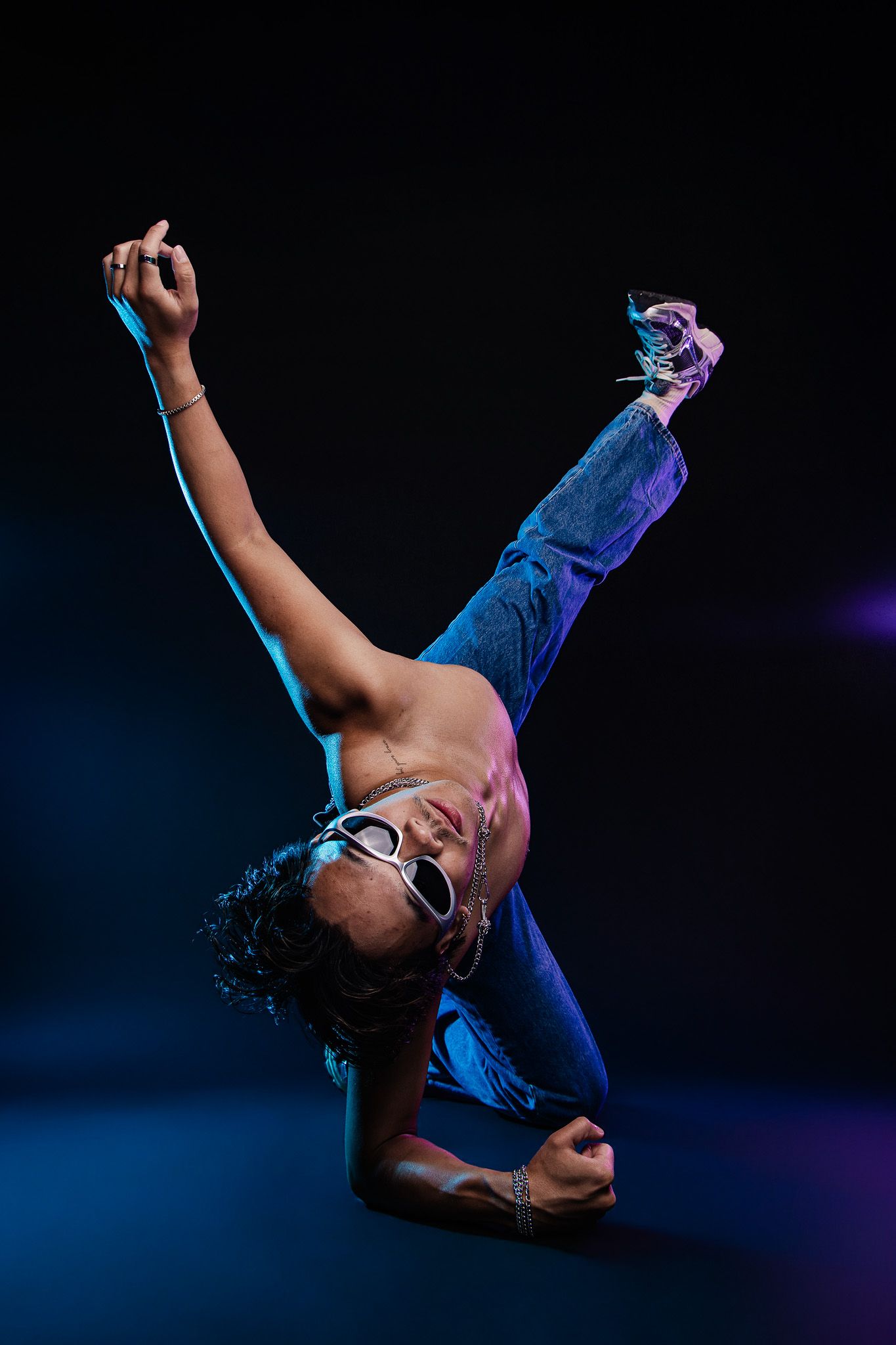 Shirtless breakdancer performing a one-handed freeze move in blue jeans and sunglasses under dramatic neon lighting.