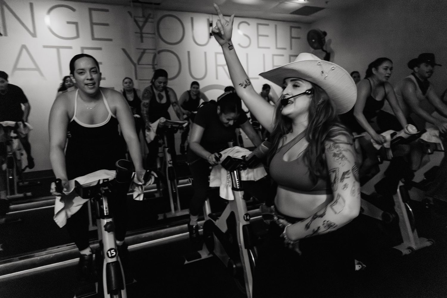Black and white photo of women energetically cycling in a spin class, one wearing a cowboy hat and raising her hand.