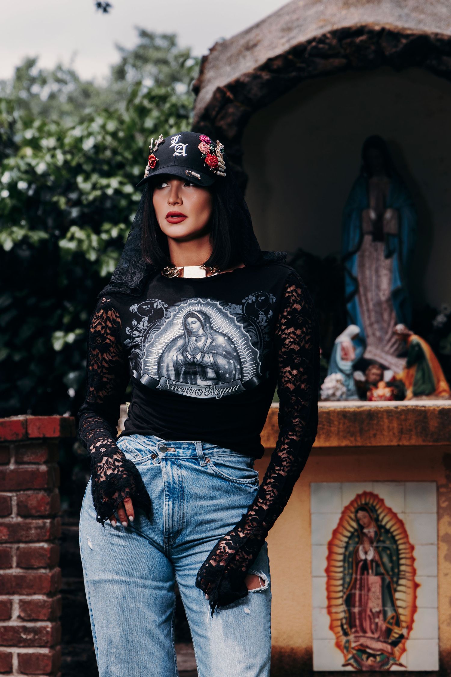 Woman in black lace top, Virgin Mary tee, and embroidered cap poses near religious shrine with blue jeans.