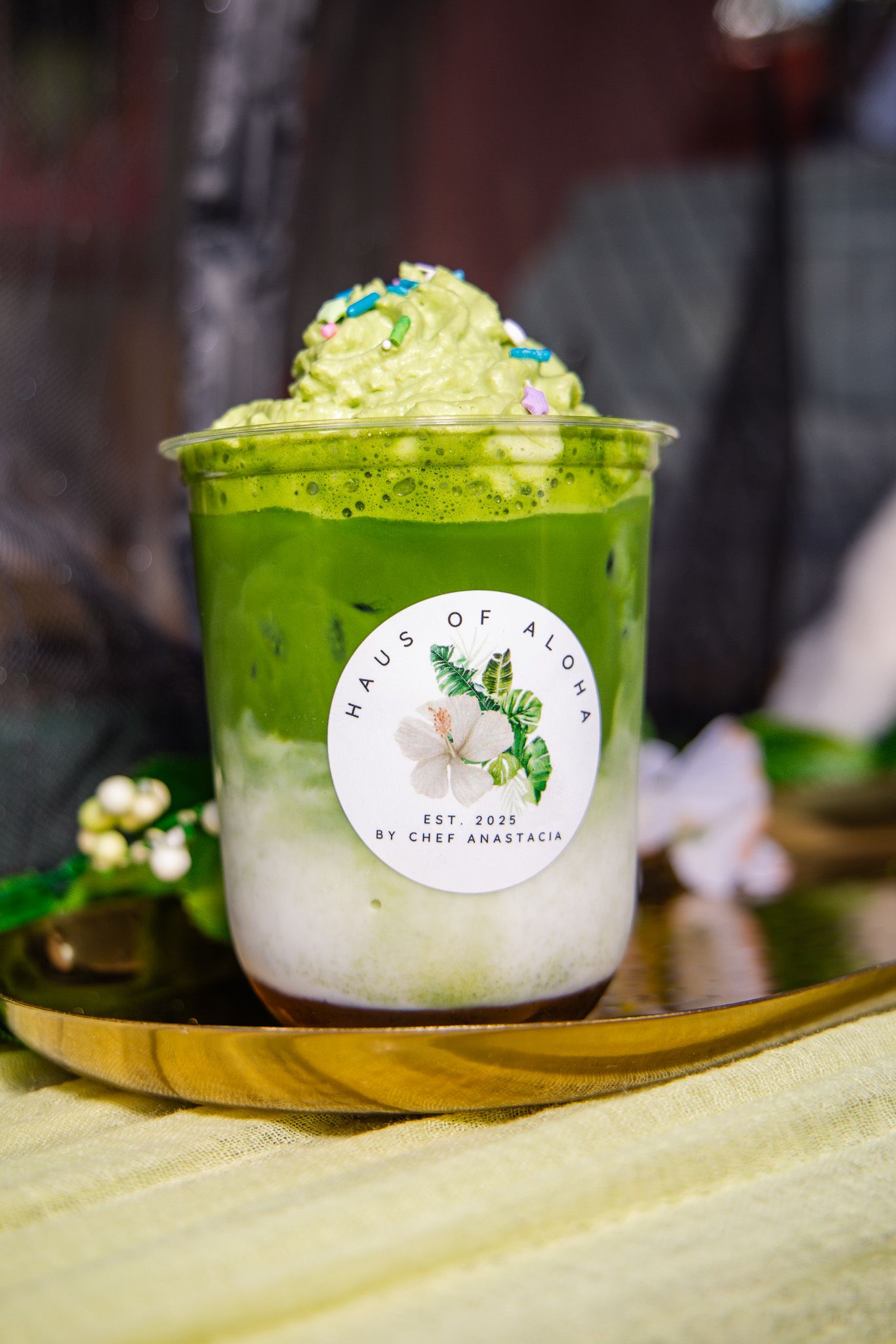 A layered matcha green tea drink with whipped cream and sprinkles in a branded cup on a gold plate.
