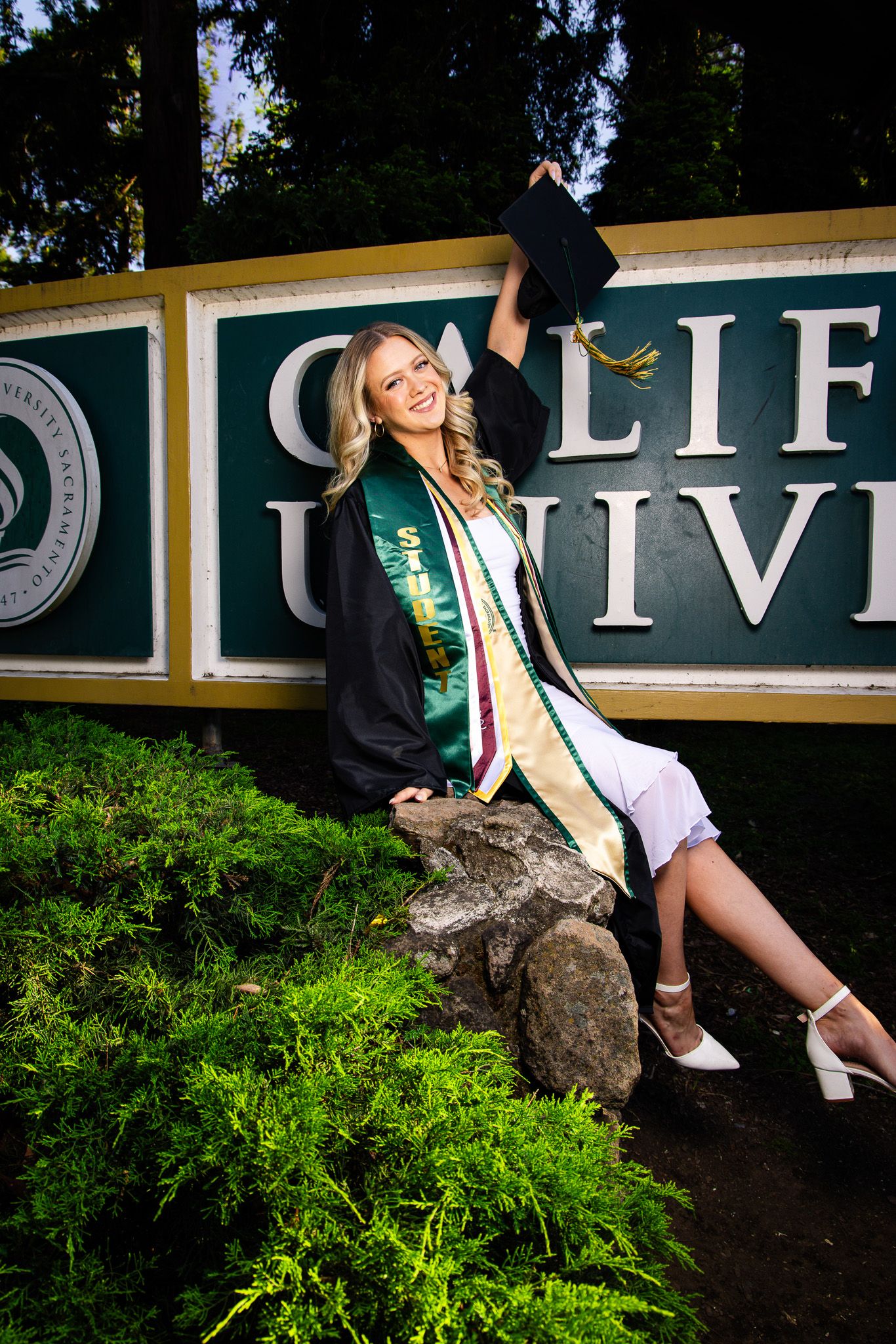Smiling female graduate in black gown and green sash poses with cap at California University sign outdoors.