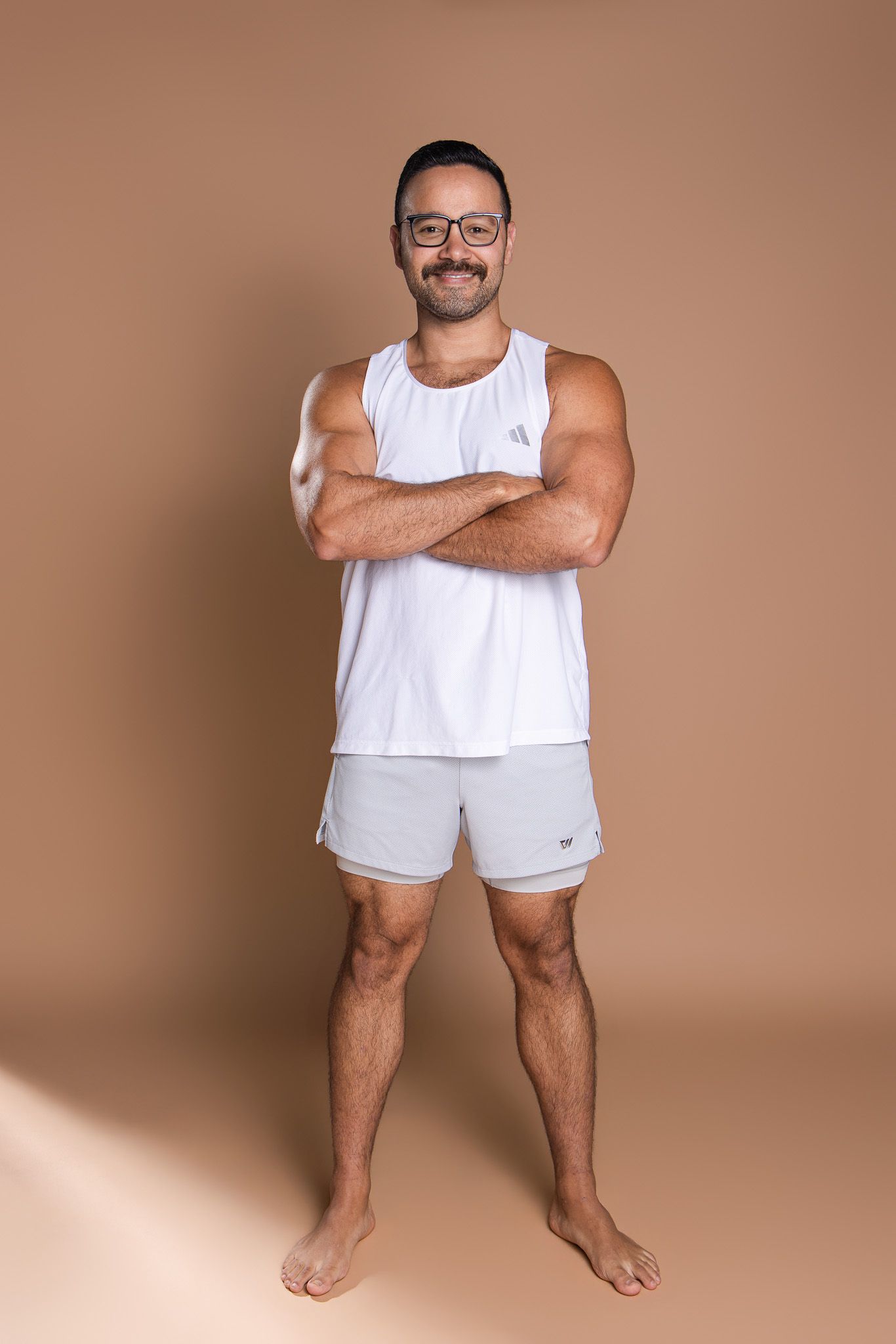 Smiling man in white tank top and gray shorts standing with arms crossed in beige studio.