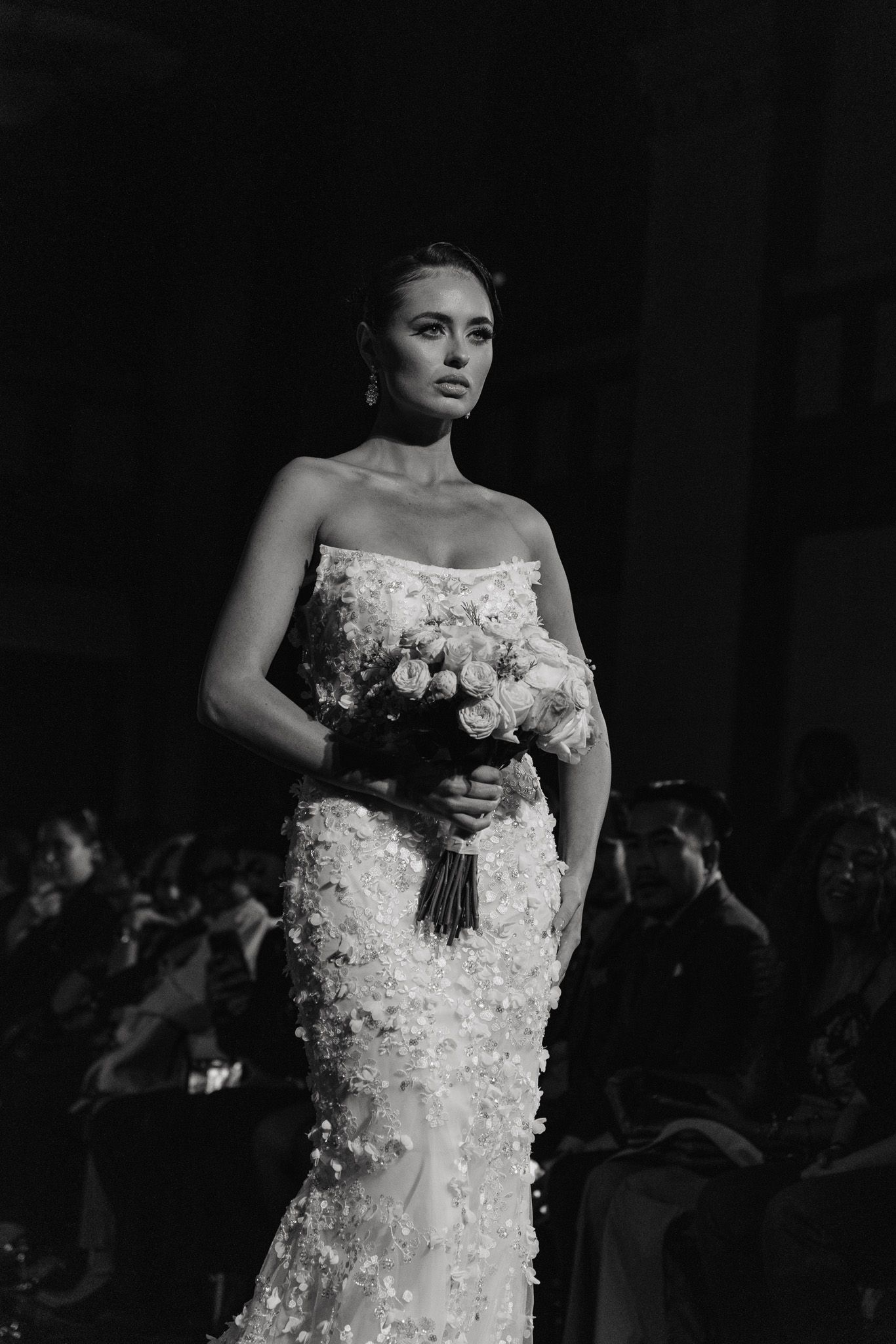 A model walks the runway in an elegant strapless lace wedding gown while holding a bouquet of flowers.