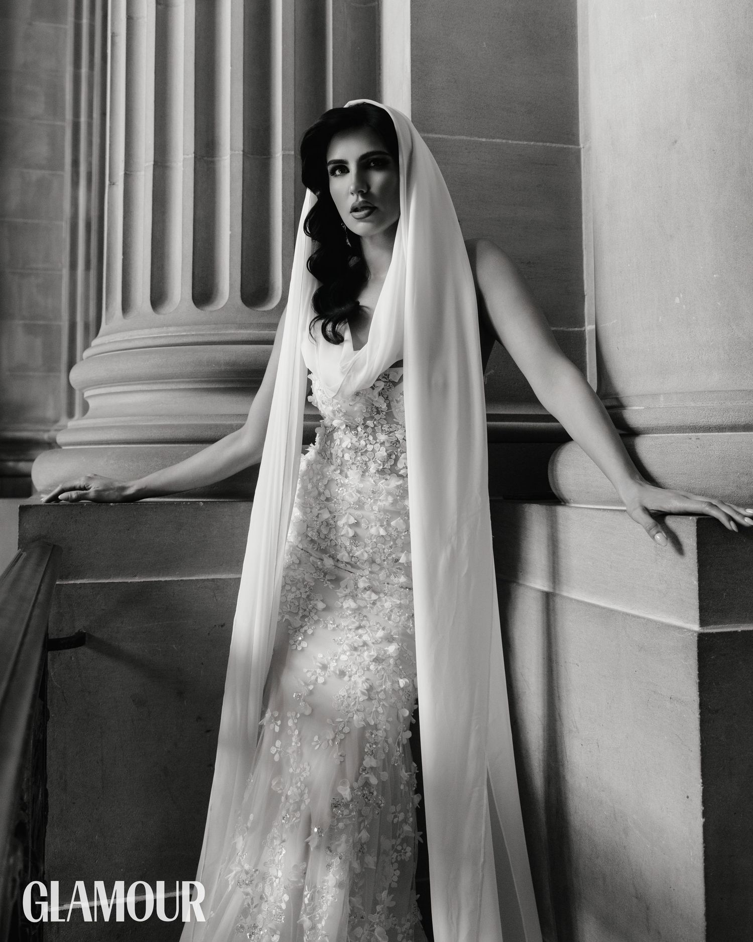 Elegant black and white bridal portrait showing a bride in lace gown and long veil posing against classical columns.