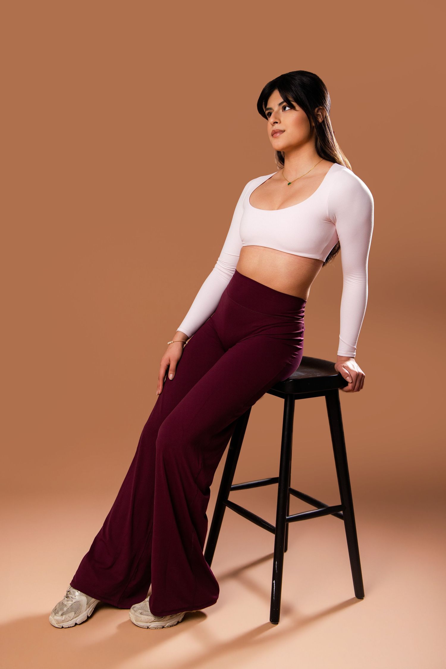 Woman in white crop top and burgundy palazzo pants poses elegantly on black chair against warm brown backdrop.