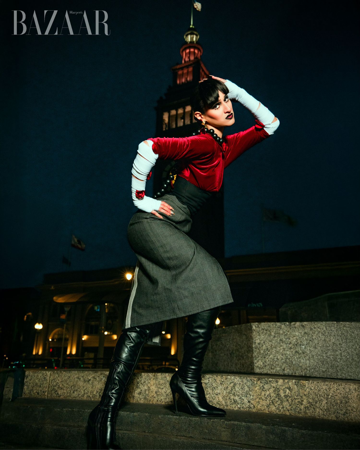 Fashion model in red sweater and gray skirt poses dramatically at night against illuminated building backdrop.