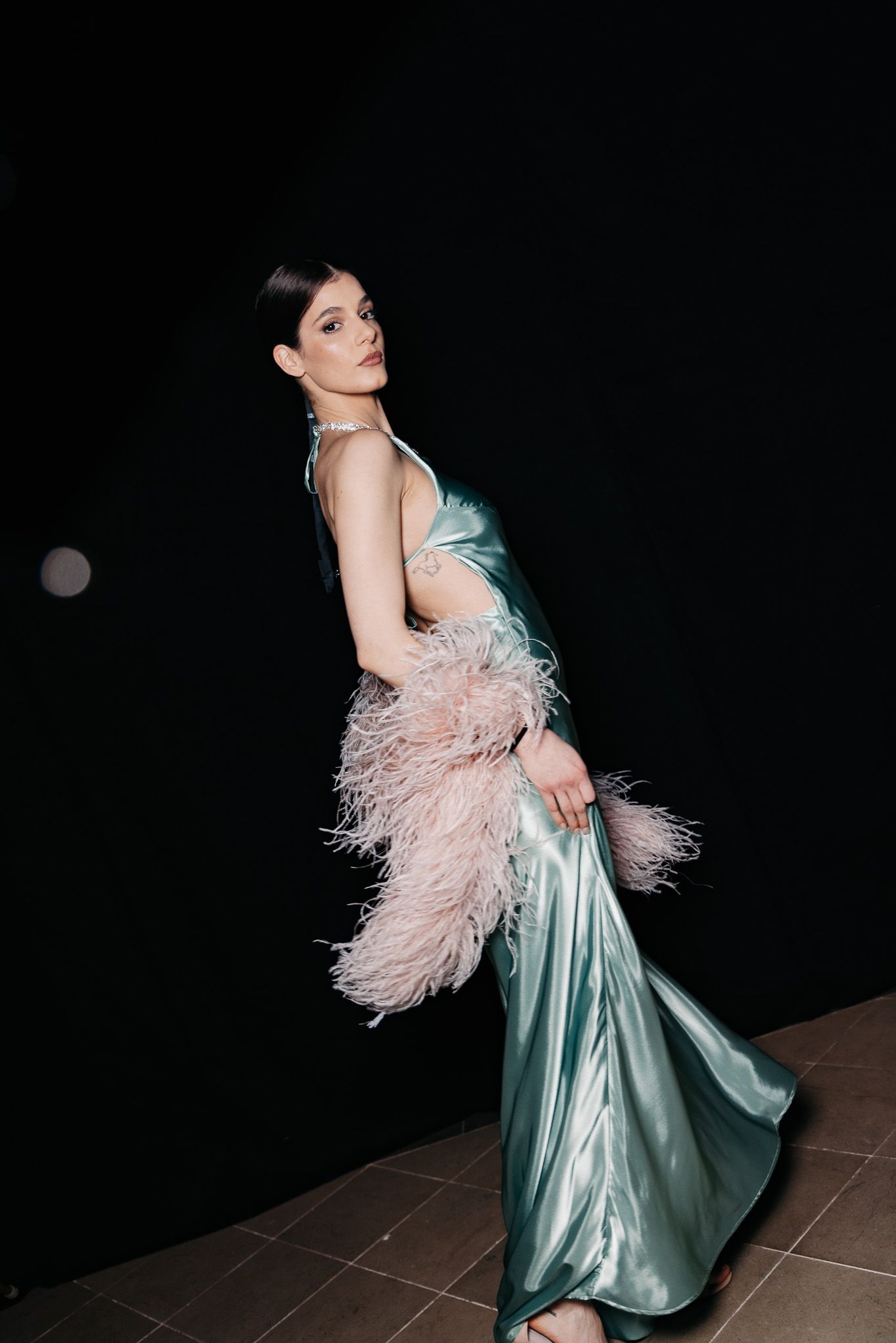 Model in flowing mint satin gown with pink feather detail poses dramatically against dark background.