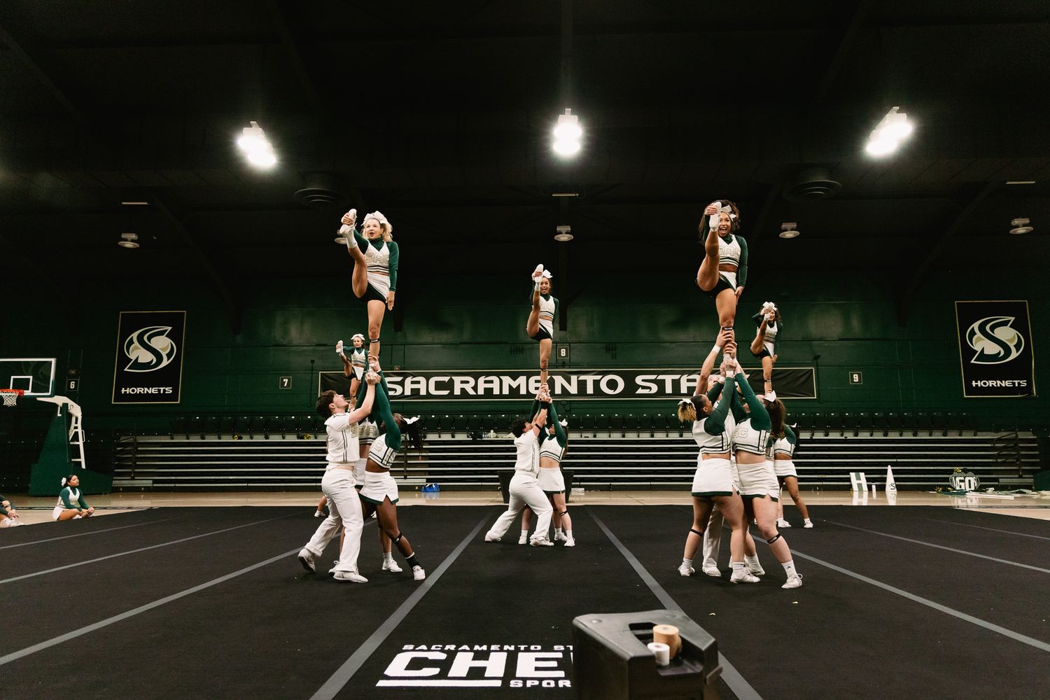 Sacramento State cheerleaders performing a pyramid stunt in a green and white gymnasium during a cheer competition.